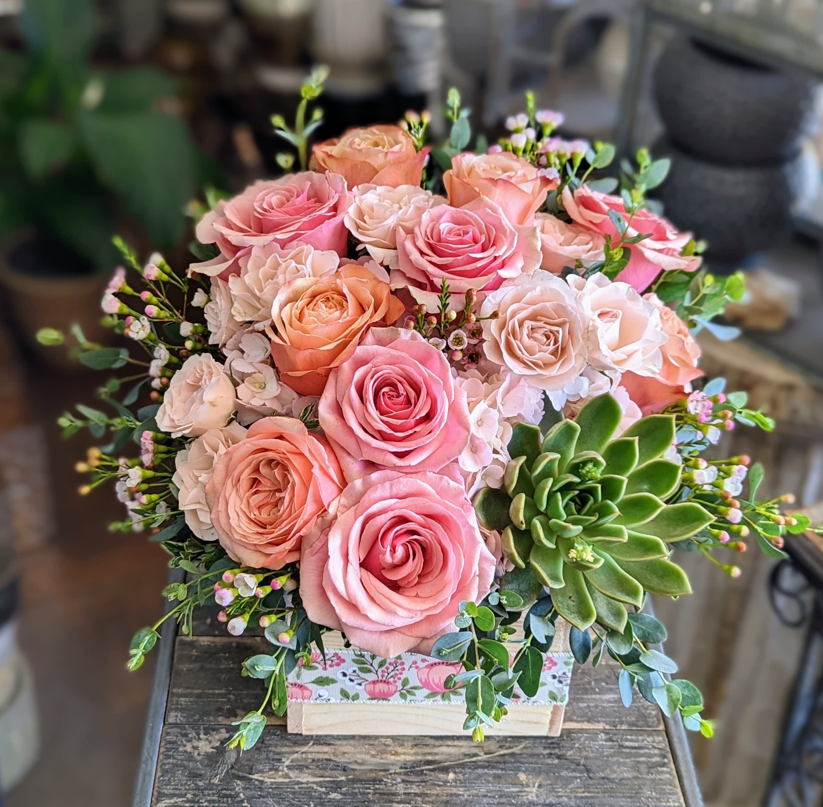 Chloe Chloe - Chloe is a tender daydream in bloom—graceful, fresh, and quietly radiant. This charming arrangement bursts with rosy delight: velvety pink and peach garden roses swirl among creamy blush spray roses and clusters of soft hydrangea, creating a harmonious palette of pastel warmth. A vibrant echeveria succulent offers a playful, modern twist—its rosette form echoing the beauty of the roses while adding a touch of grounded elegance. Sprigs of waxflower and fresh greenery weave through the design, adding texture and lightness. Presented in a wooden box adorned with delicate floral print, Chloe feels like a keepsake from a secret garden—perfect for celebrating love, friendship, or simply brightening someone's day. Available exclusively from Windmill Floral Expressions—with delivery throughout Las Vegas and Henderson—where timeless beauty meets heartfelt design.