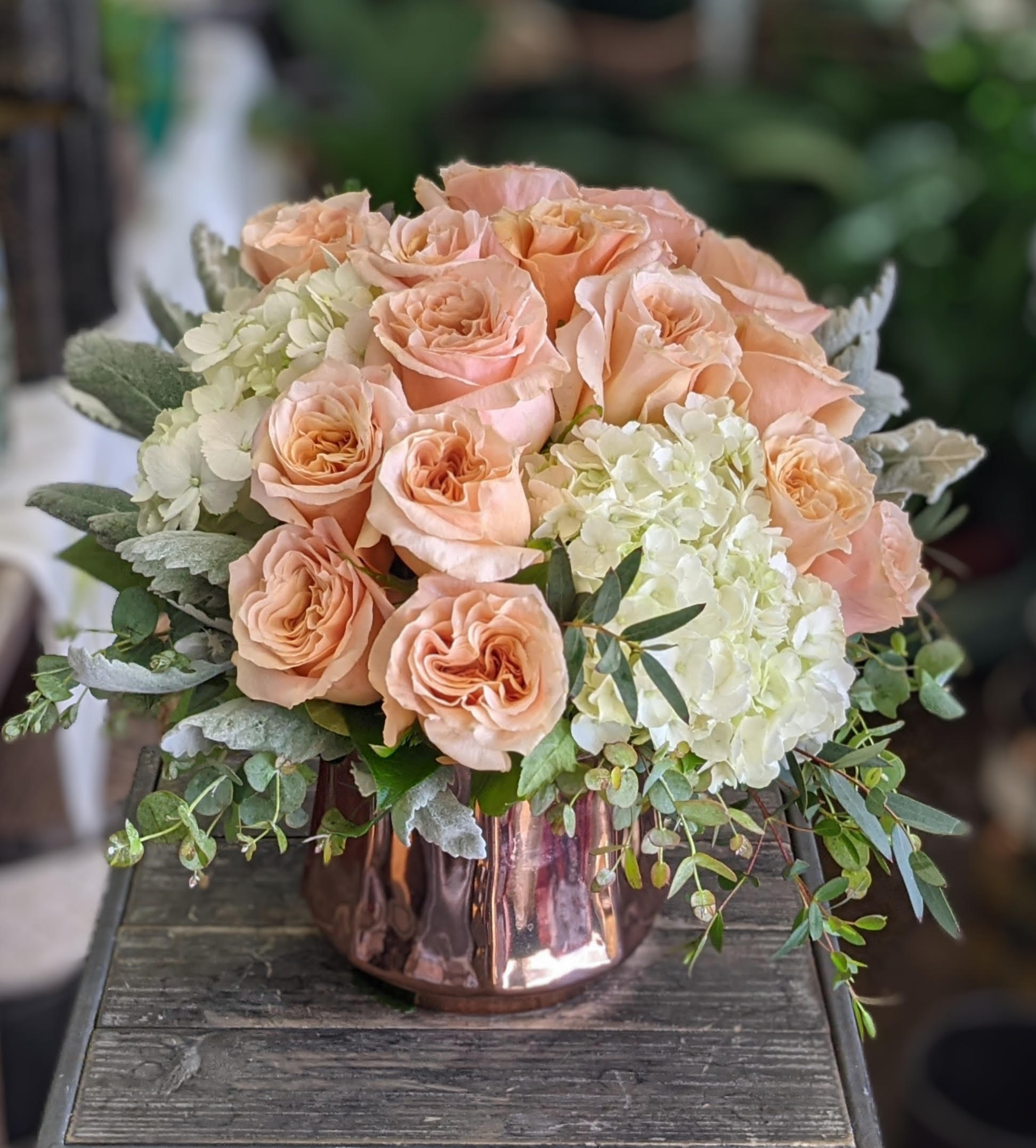 Bianca Bianca - Bianca is a graceful celebration of quiet elegance and refined romance. At its heart, fifteen peach roses bloom with soft, ruffled petals that radiate a gentle apricot glow. Billowy white hydrangeas lend an airy sophistication, their cloud-like clusters offering a soft contrast in both texture and tone. Silvery dusty miller leaves, velvety and cool, weave through the design, adding a subtle shimmer that harmonizes with the blush palette. Cascading sprigs of eucalyptus and fresh greenery spill gently over the edges, evoking a sense of natural movement and unstudied beauty. Anchored in a sleek rose-gold metallic vase, the arrangement reflects light with a modern, luminous charm—perfectly balancing softness with style. Romantic, refined, and effortlessly serene, Bianca is a floral whisper that speaks volumes. Available for delivery in Las Vegas and Henderson from Windmill Floral Expressions, where artistry and emotion bloom in perfect harmony.
