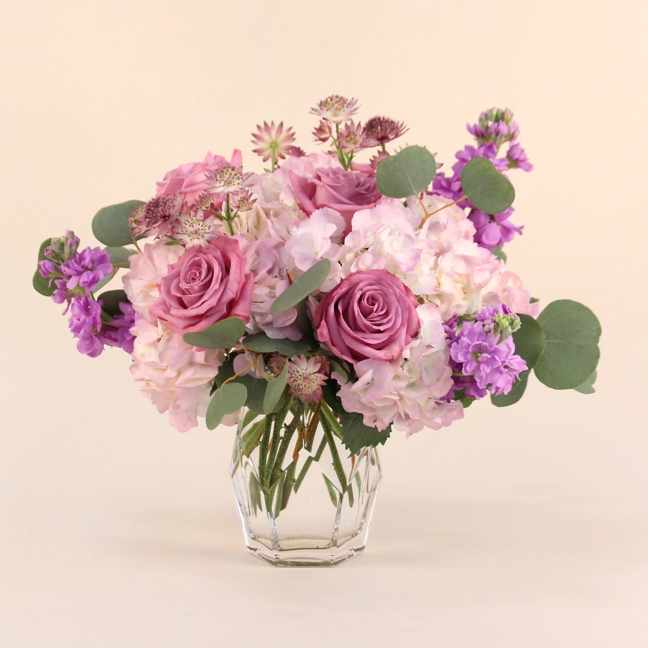Sugar Plum - A lush arrangements of lavender and mauve flowers in a clear cylinder vase. Flowers may include roses, hydrangeas, astrantia, stock and silver dollar eucalyptus.  Vase dimension: 5” x 8&quot;