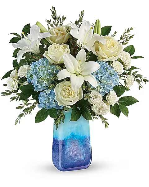 Teleflora's Ocean Sparkle Bouquet - Like a calm morning by the sea, this beautiful bouquet of blue and white blooms arranged in a stunning, hand-blown art glass keepsake vase, invigorates and inspires.  This beautiful bouquet includes blue hydrangea, white roses, white spray roses, parvifolia eucalyptus, and lemon leaf. Delivered in a Blue Lapis Gemstone Art Glass vase.
