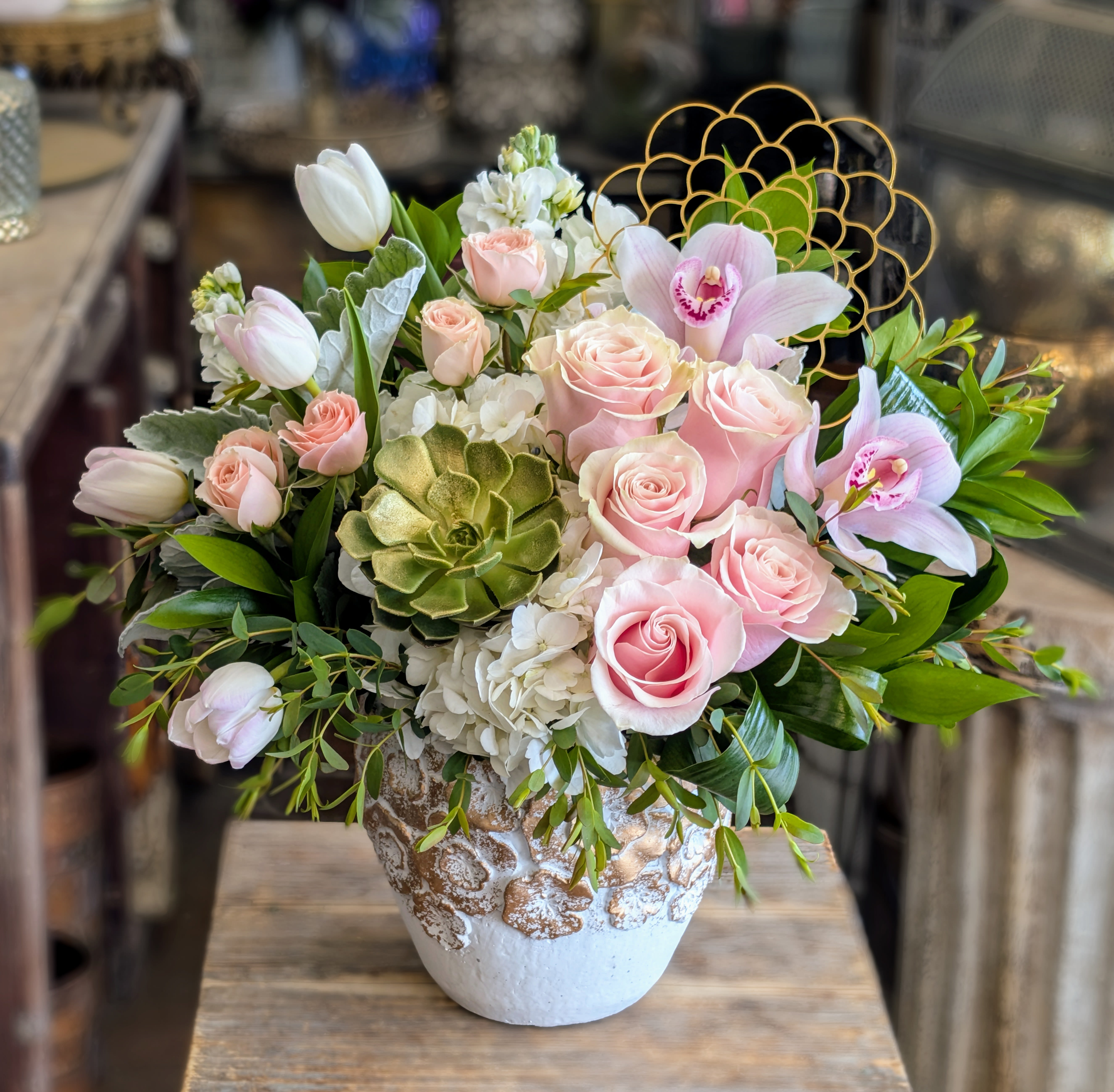 Charlotte - Charlotte is the embodiment of timeless grace and gentle beauty. A romantic palette of blush pink roses, delicate tulips, and soft white hydrangeas evokes the charm of an English garden at first light. Pale cymbidium orchids add an elegant flourish, while a sculpted green succulent at the heart brings unexpected texture and a hint of modernity. Wisps of dusty miller,  lush greenery, and a golden geometric accent complete the design, adding softness and shimmer in perfect balance.  Cradled in an artisan ceramic vessel adorned with gilded coral-like texture, Charlotte feels both refined and whimsical—a perfect gift for birthdays, baby showers, or simply to brighten someone’s world. Now available for delivery in Las Vegas and Henderson from Windmill Floral Expressions.  