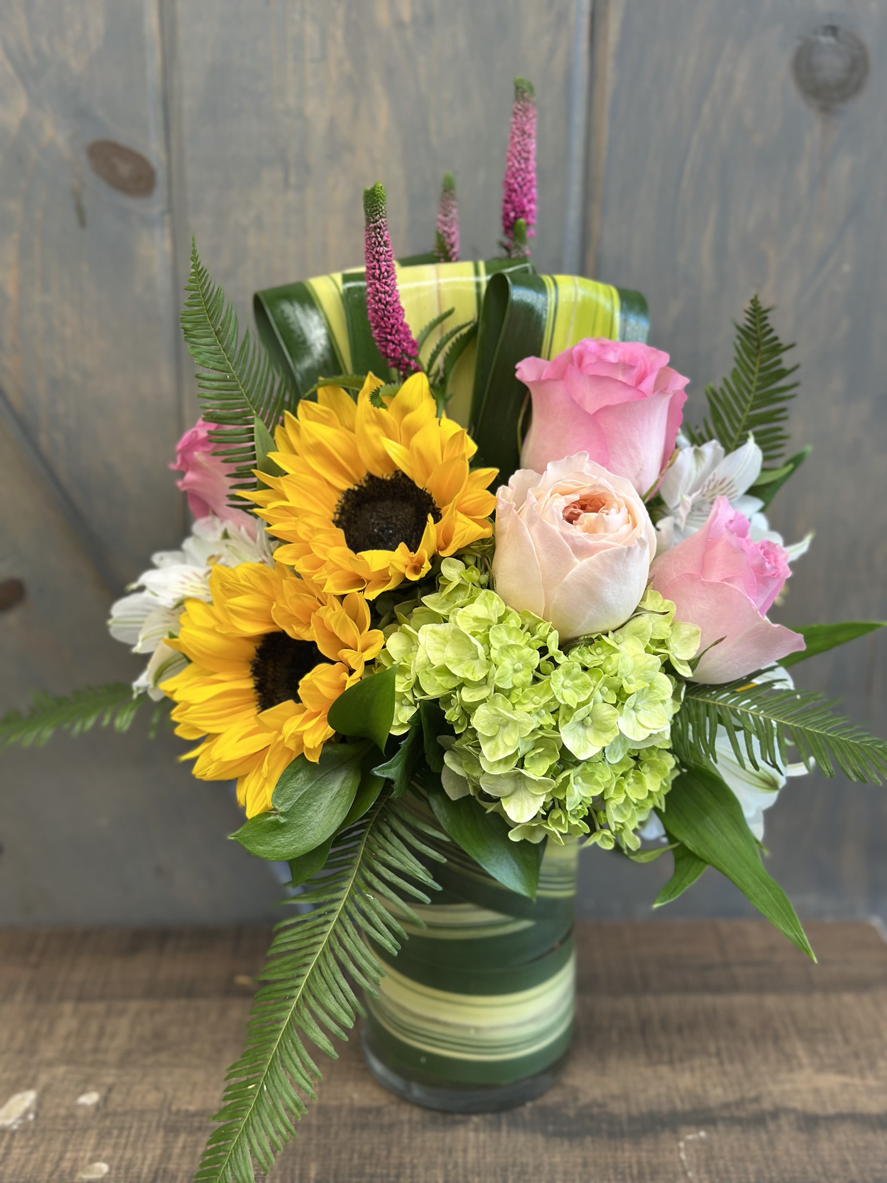 Summer Fun - The perfect arrangement to  lift spirits no matter the occasion. The flowers included in this design are Sunflowers, Roses, Garden Roses, Mini Hydrangea, Veronica, Alstroemeria, Umbrella fern, and Flax leaf