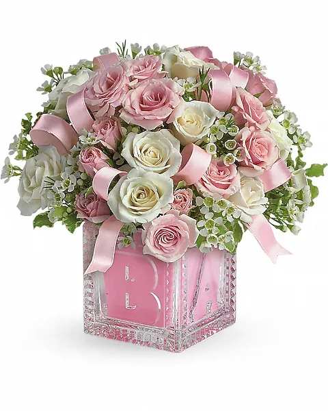 Baby's First Block by Teleflora - Pink - Celebrate the cutest baby girl on the block's arrival with this charming glass baby block that arrives chock full of pretty flowers. Perfect for baby showers, too! The glass block will make an adorable display piece for years!  Pretty light pink roses, pink and white spray roses, miniature gerberas and waxflower and pink satin ribbon are delivered in a clear baby block with a pink liner. One amazing delivery deserves another, don't you think?  