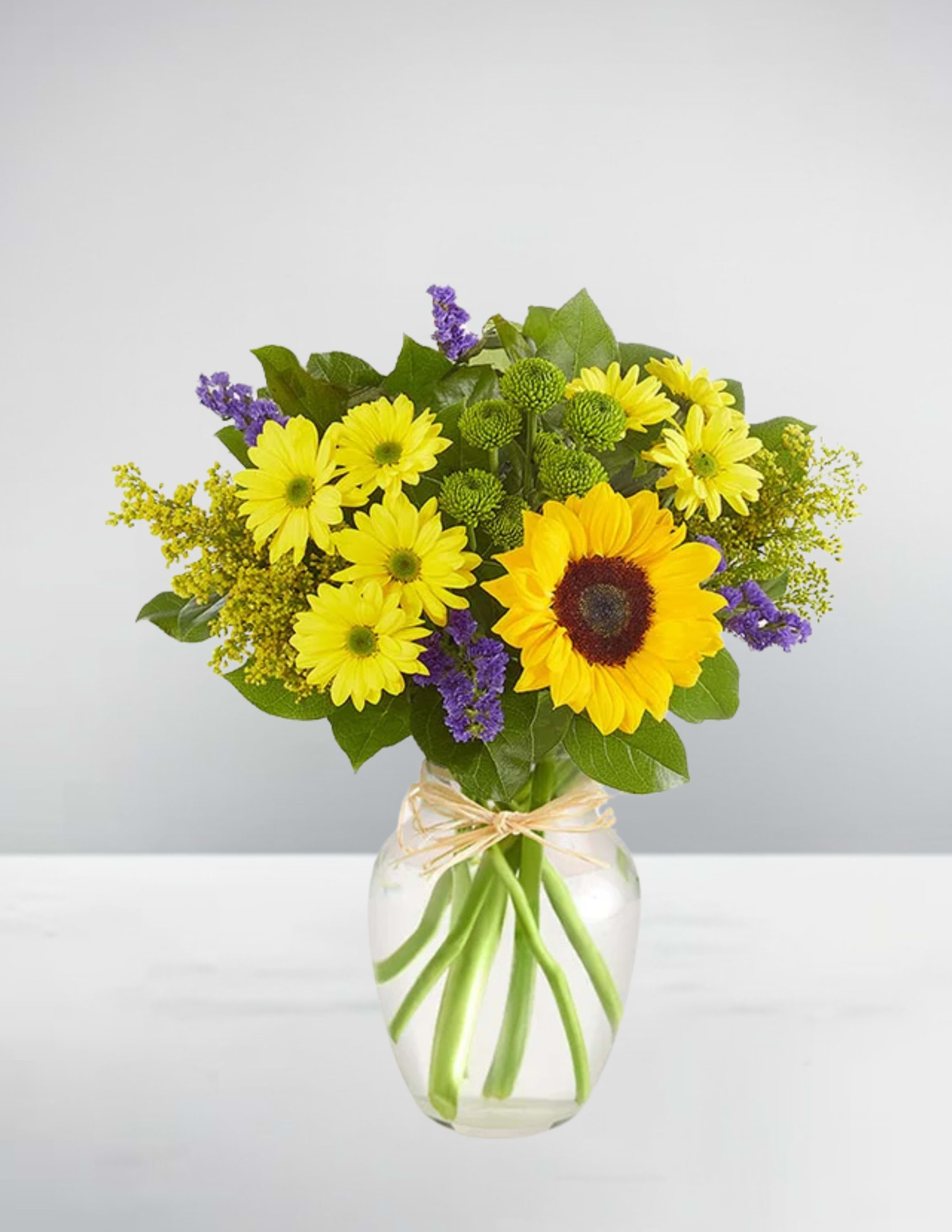Sunshine meadow - Sunshine Meadow Let happiness bloom with Sunshine Meadow, a vibrant and uplifting bouquet designed to bring a burst of joy to any room. This cheerful arrangement features a radiant sunflower as the focal point, surrounded by sunny yellow daisies, playful green button poms, and touches of purple statice and golden solidago. Hand-arranged in a classic clear glass vase tied with rustic raffia, this bouquet captures the essence of a fresh summer meadow. Perfect for birthdays, thank-yous, get-well wishes, or just because—Sunshine Meadow is a thoughtful way to brighten someone’s day!