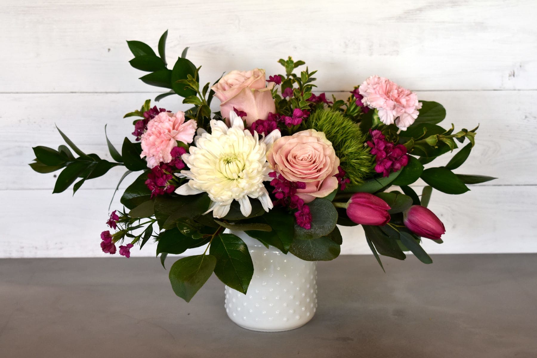 Kristine  - This arrangement features a spring mix of pink roses, pink carnations, chrysanthemums, tulips, greens, and filler in a white hobnail vase. Please be advised, that the flowers and vase may vary.