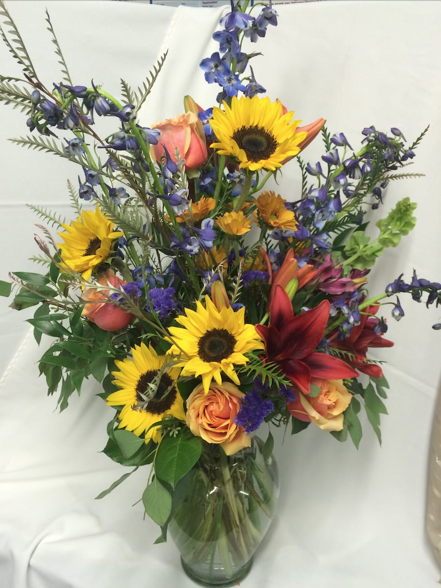 Radiant Sunflower Bouquet - Sunflowers enhance this beautiful mix of delphinium, scarlet lilies and orange roses Additional flowers may vary depending on selection available at time of order. Vase design will vary based upon availability at time of delivery