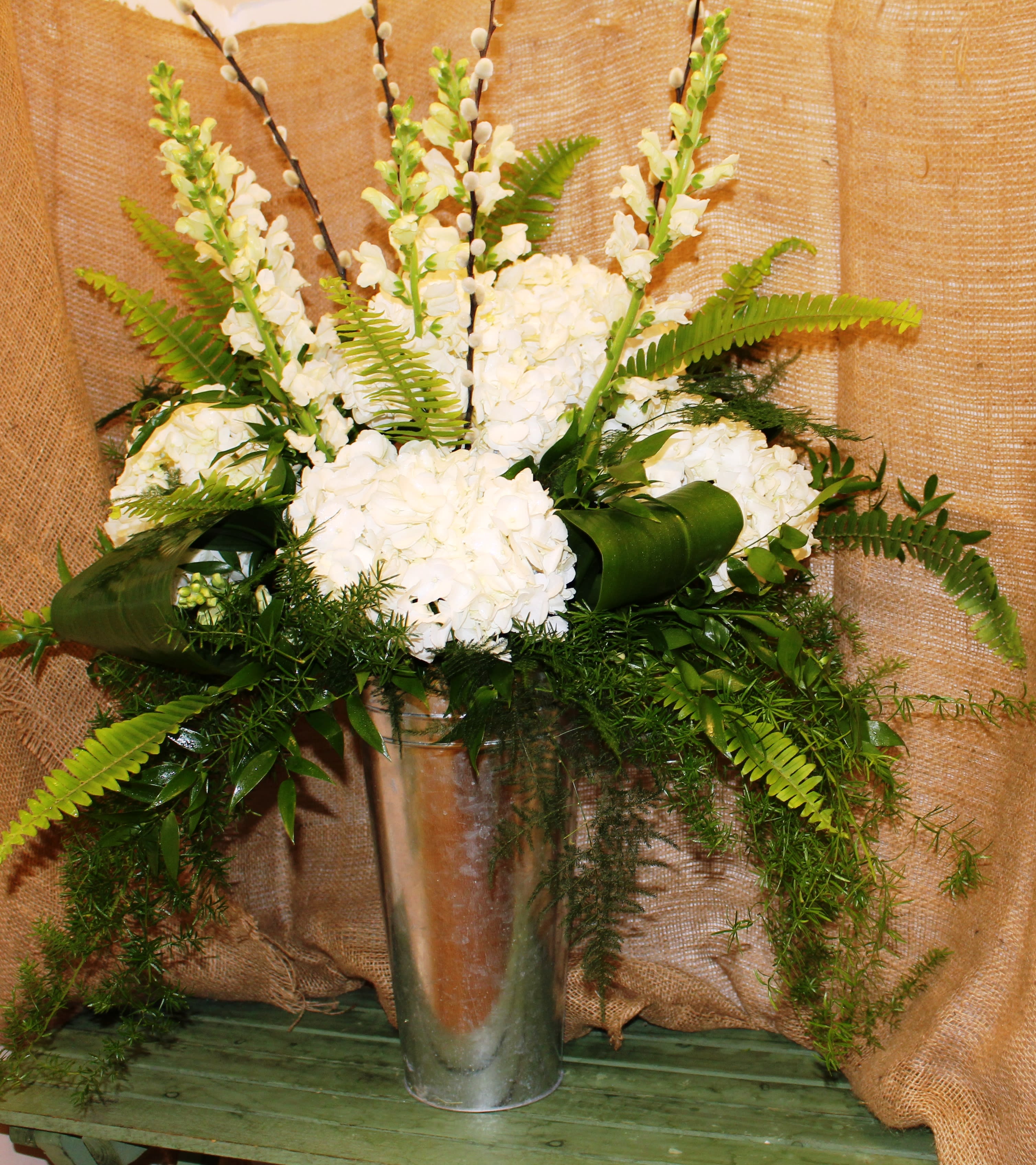 Serene Reflections - Commemorate a life well-lived with this beautiful standing piece. A galvanized pail filled with a fresh mix of flowers in a stunning white and green color line. Featuring multiple types of greenery, large white hydrangea, and pussy willows, this arrangement is absolutely stunning. Can be sent alone, or would look great in pairs anchoring the casket or urn to send off a loved one. 
