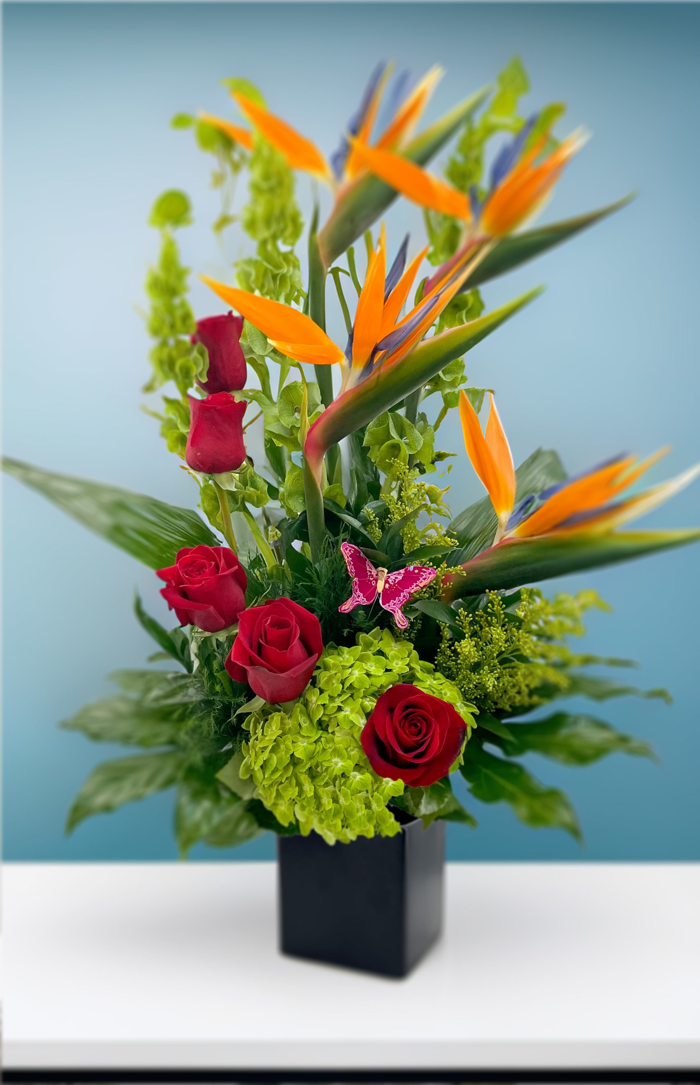Tropical Romance - The tropical region is renowned for its stunning array of unique and vibrant flowers. In this sophisticated arrangement, we showcase the beauty of tropical bird of paradise flowers alongside elegant red roses and lush green hydrangeas.
