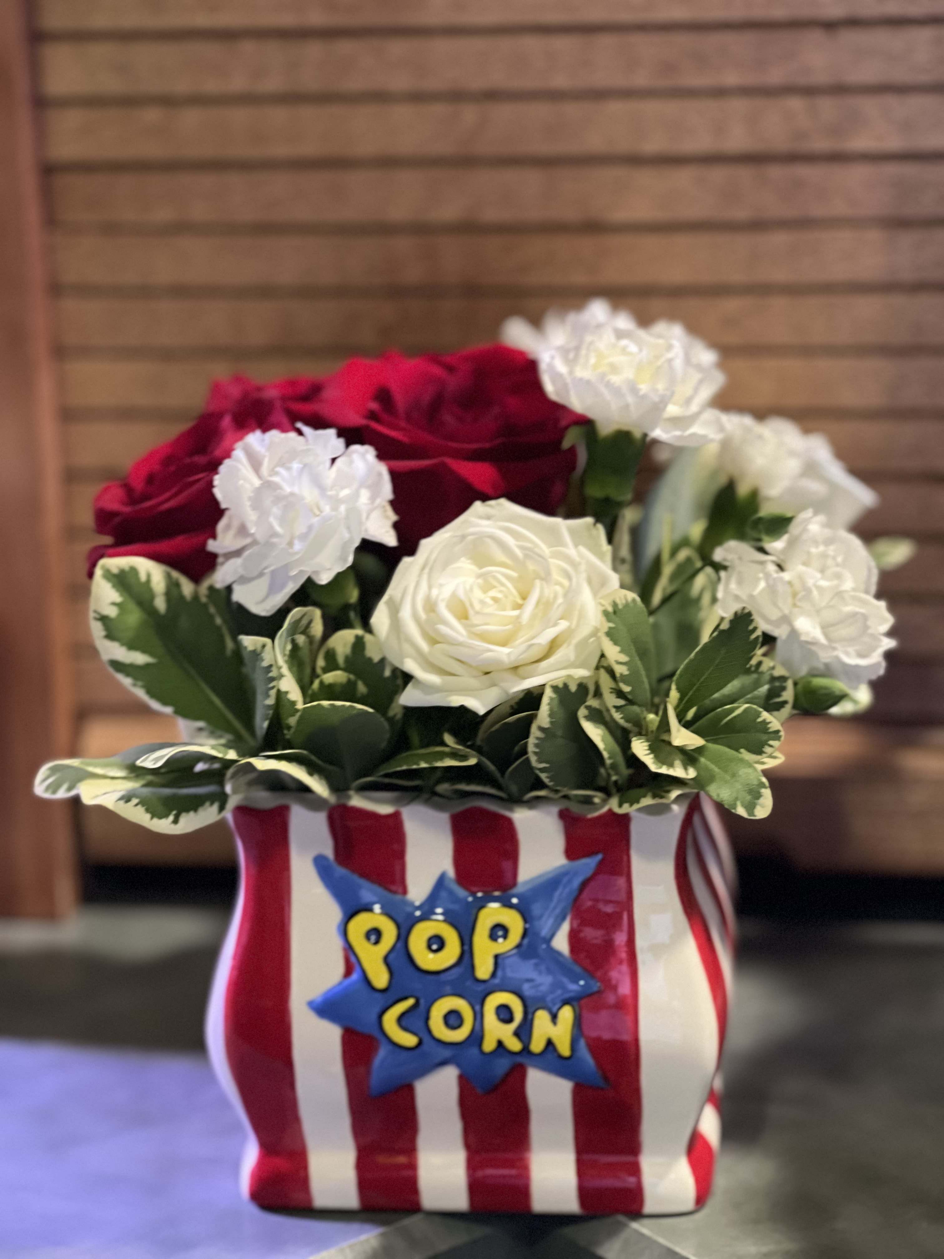 Movie Night Movie Night - What better to celebrate TV night in American than this cute little bag of red and white stripes with the word popcorn created with a few Red Roses, white mini carnations, (to mimic popcorn) green trick dianthus and variegated Pittosporum. The ceramic container can be used after the flowers have faded to house your remote or gaming controllers. Inventory count: 3