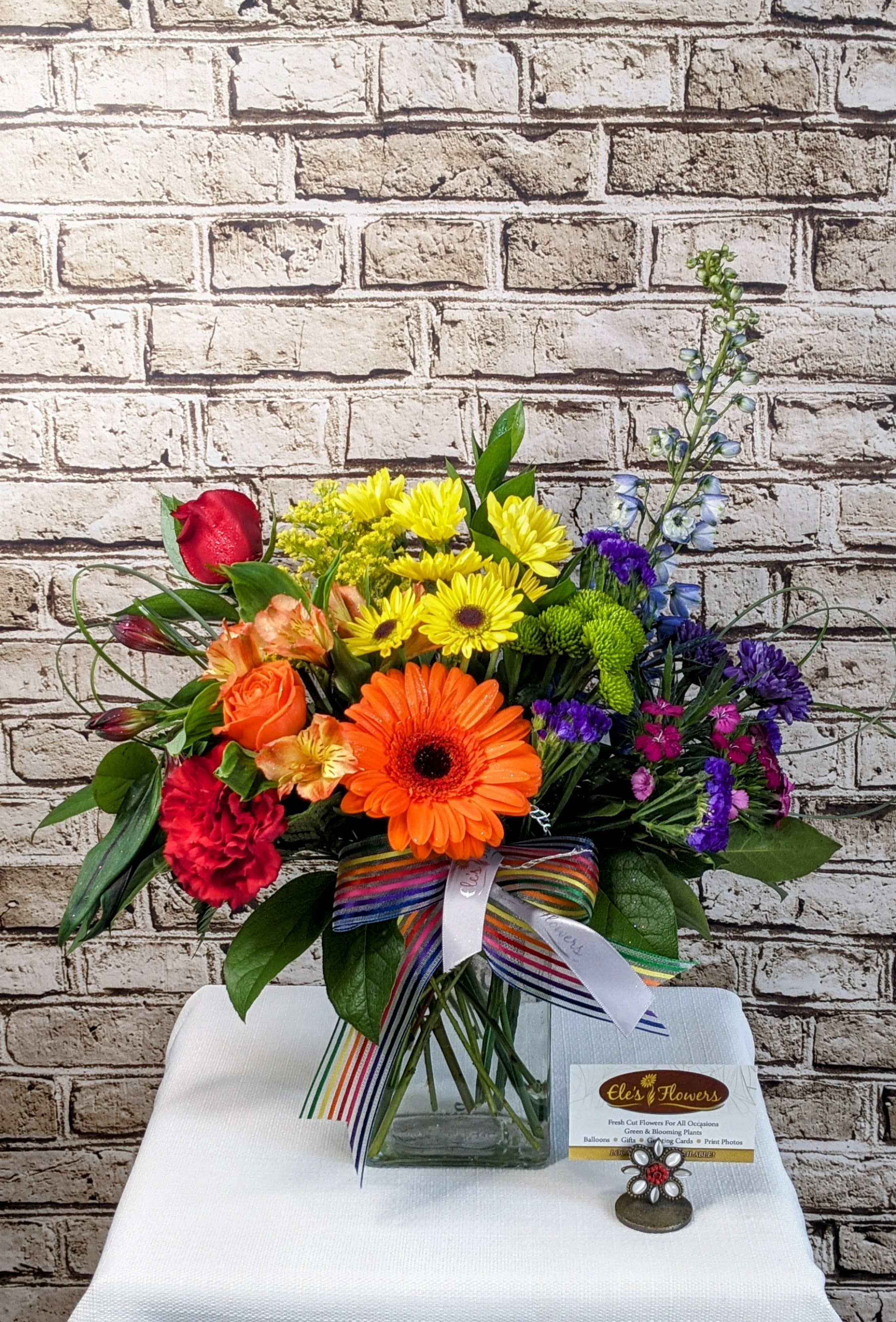 Prism Blooms   - A radiant celebration of color, Prism Blooms is a joyful expression of nature’s full spectrum. Arranged in a clear glass vase, this striking design features a curated mix of fresh flowers in a vivid rainbow palette—red, orange, yellow, green, blue, indigo, and violet—layered to create a true floral prism. Every stem is chosen for its bold hue and unique texture, making this arrangement as artful as it is uplifting.  Perfect for birthdays, celebrations, pride, or simply adding a burst of happiness to any space  Approximate Dimensions: 13&quot;H x 15&quot;W