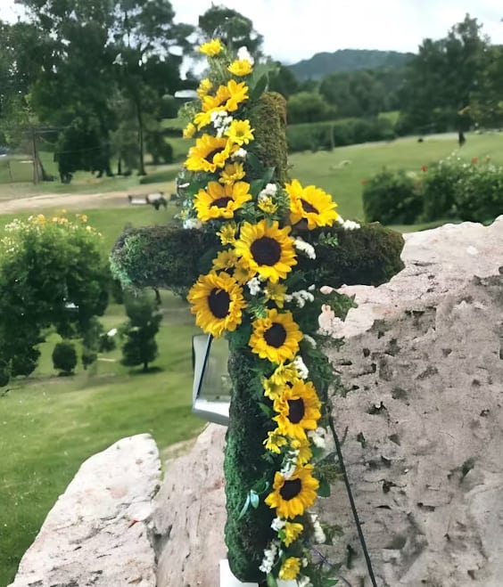 Sunflower Moss Cross Sunflower Moss Cross - A beautiful mixture of flowers and texture with artistic details. Standing at 30 inches, this moss-covered cross brings together the joyous brightness of sunflowers with the verdant texture of moss, creating a piece that is both striking and serene. Embracing the cross, the sunflowers stand tall and proud, their vivid yellow petals radiating warmth and light. These blooms are a testament to the strength and positivity that sunflowers represent, making this floral cross a visual delight and a beacon of hope and comfort. Accompanying the sunflowers are carefully selected fillers that add depth and contrast to the arrangement. These delicate blooms and greens weave through the moss, enhancing the naturalistic appeal and lending the piece an artful complexity. Displayed on an easel, the Sunflower Moss Cross is an impressive tribute for services and memorials or as a statement piece for events. It captures the eye and the heart, symbolizing eternal life and nature's continuous renewal. This piece is more than just flowers; it's a loving embrace made visible, a celebration of life through the art of floristry.