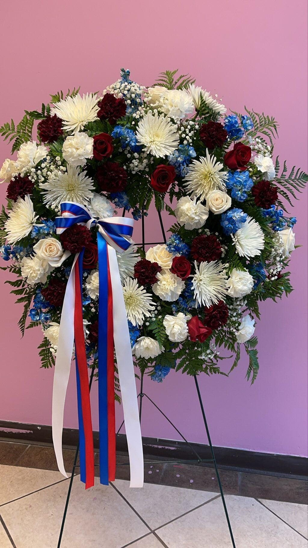 Never Forgotten - Honor to whom honor is due through this sympathy wreath. This wreath contains red roses, carnations, white spider mums, blue hydrangea. Also includes red, white and blue ribbon (as shown).