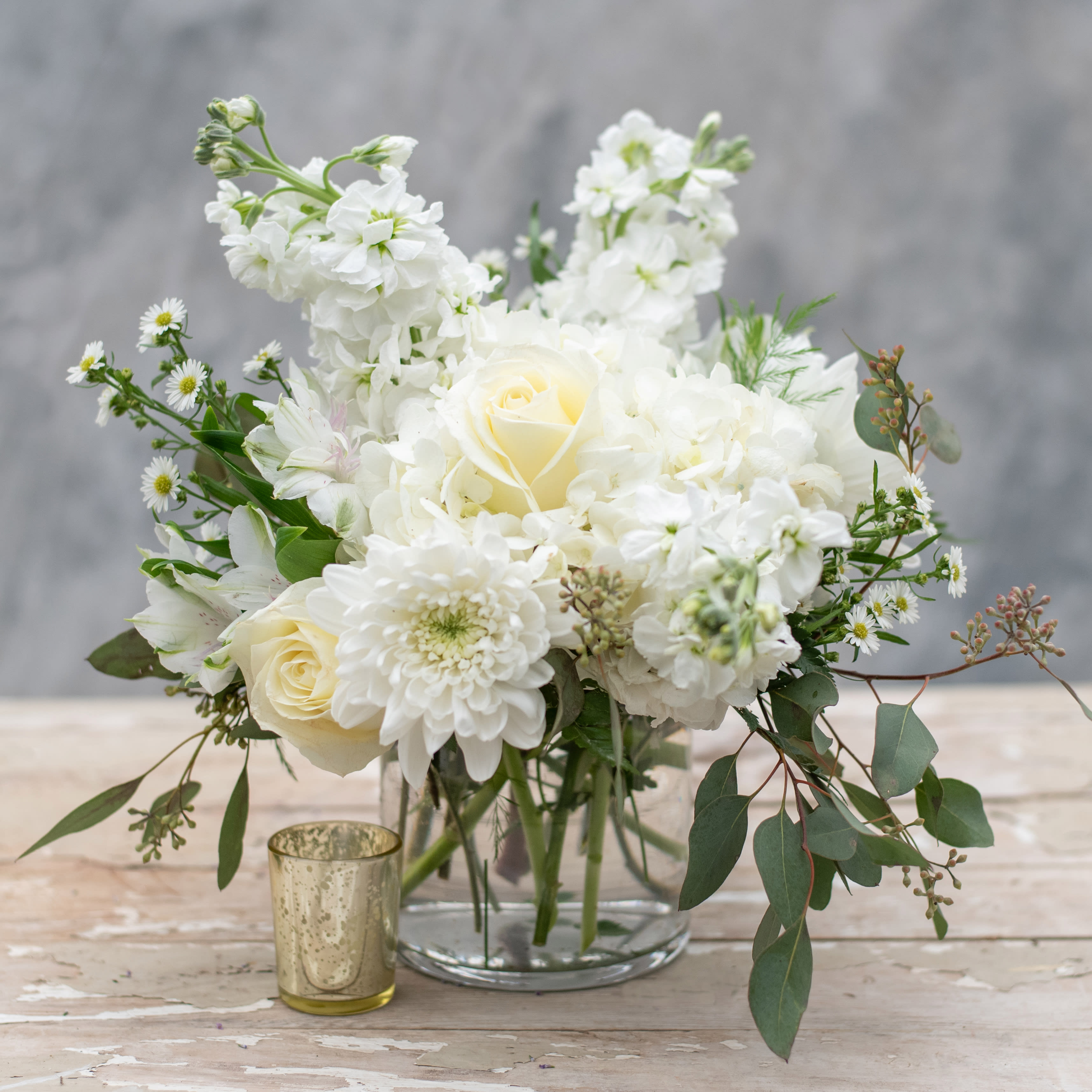 Pearly White - An elegant statement for any table in your home.  This 5&quot; glass cylinder vase is bursting with white flowers, including hydrangea, stock, chrysanthemums, roses and accented with monte casino and premium greens.