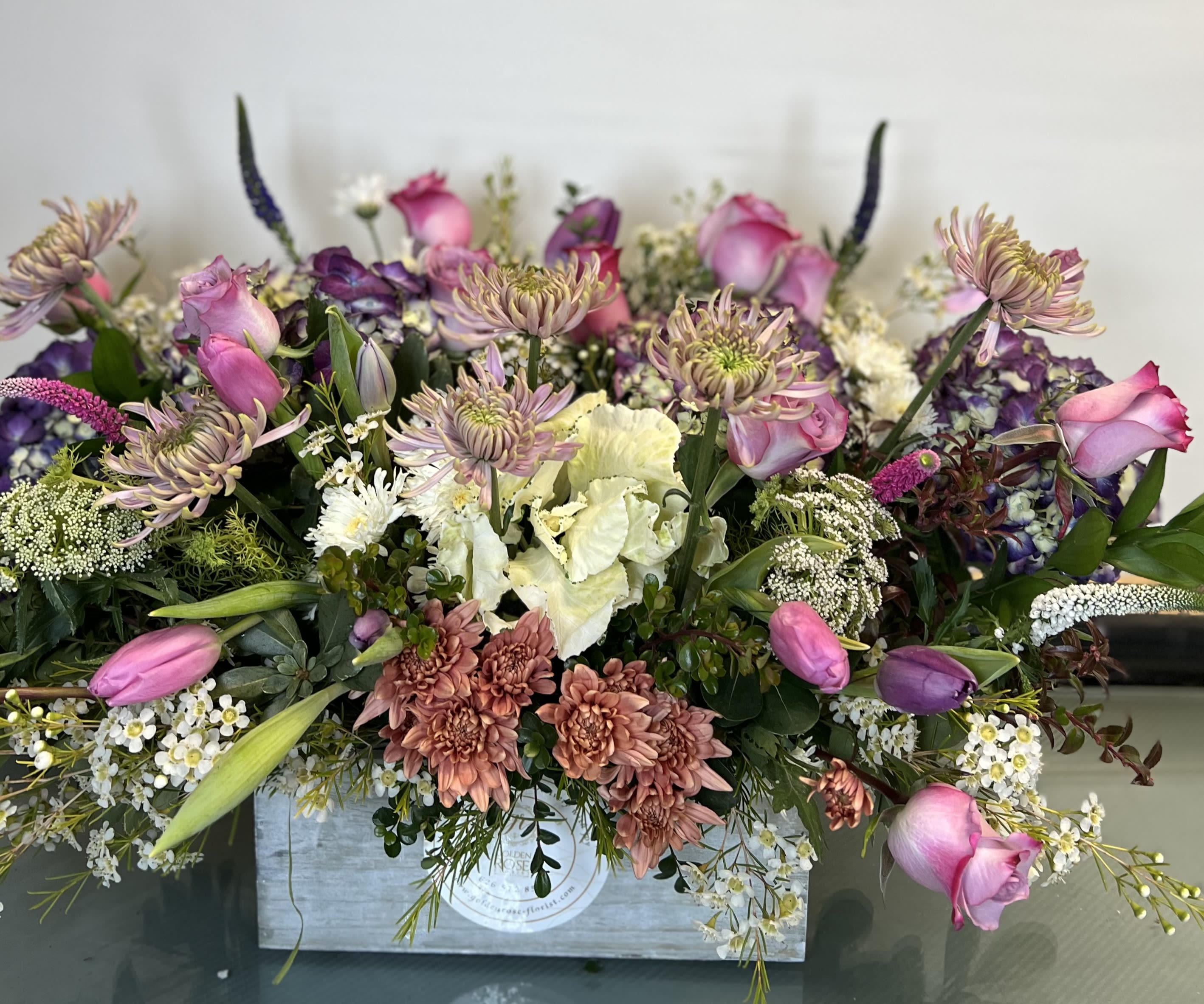 Lavender Garden Box - A stunning floral design in a rustic wooden box featuring soft pink roses, lavender tulips, spider mums, hydrangeas, and seasonal fillers in rich purple, mauve, blush, and ivory tones. This wild-style garden arrangement brings elegance with a touch of whimsy — perfect for birthdays, get well wishes, sympathy, or simply to brighten someone’s week.  Please note: Flower substitutions may occur based on seasonal availability or what we have in stock, but we will always maintain the overall look, color palette, and quality of the design.