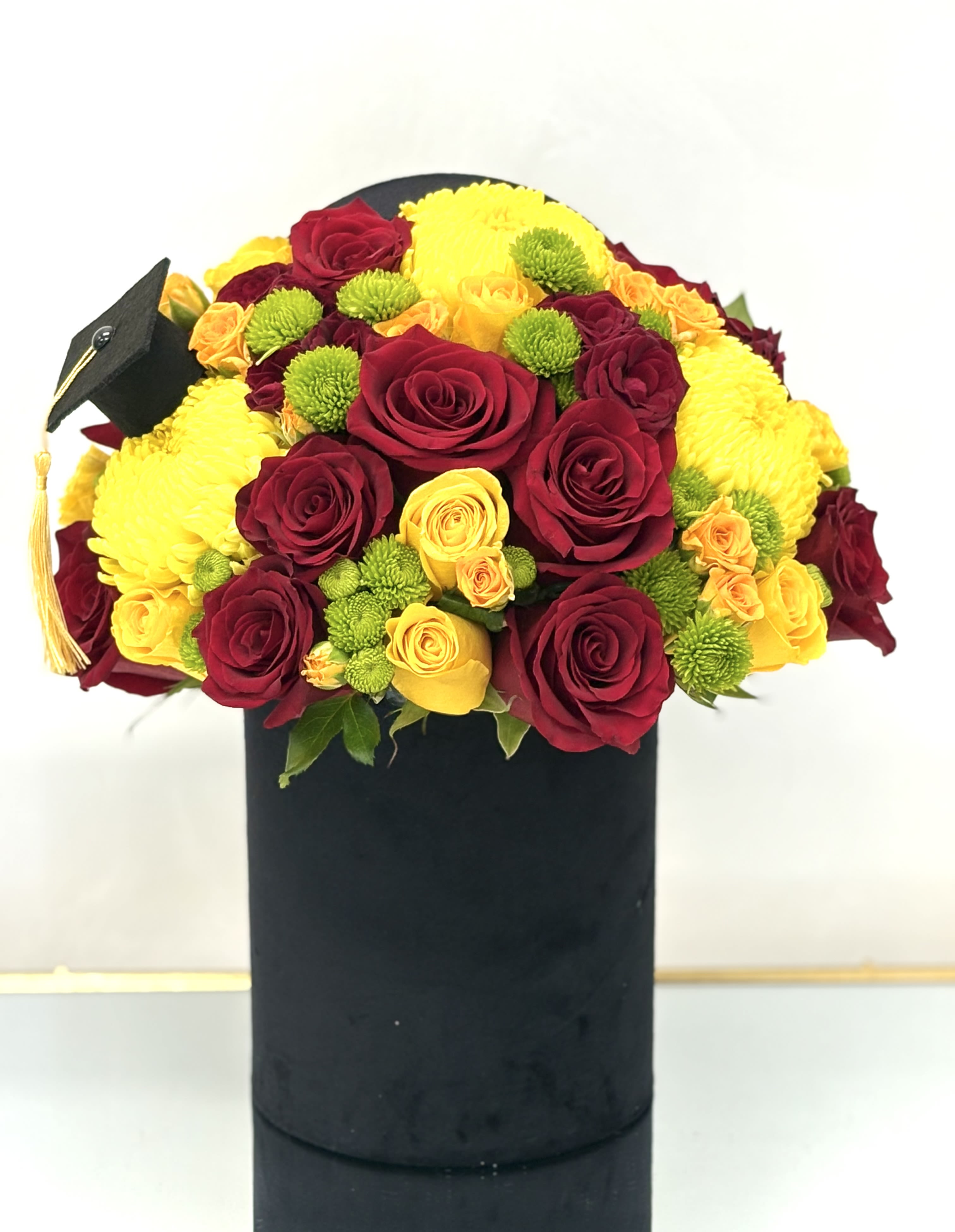 Velvet Diploma Hat Box  By VLM Flowers - Celebrate academic achievement with timeless elegance. This sophisticated arrangement features a luxurious mix of deep red and vibrant yellow roses, golden-hued chrysanthemums, and playful green accents, all artfully arranged in a sleek black velvet hatbox.   