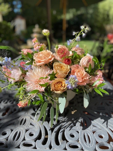 Peaches and Pastels - a selection of peach toned blooms that includes roses, stock, and renunculus with other seasonal pastel flowers arranged in a white ceramic container. 