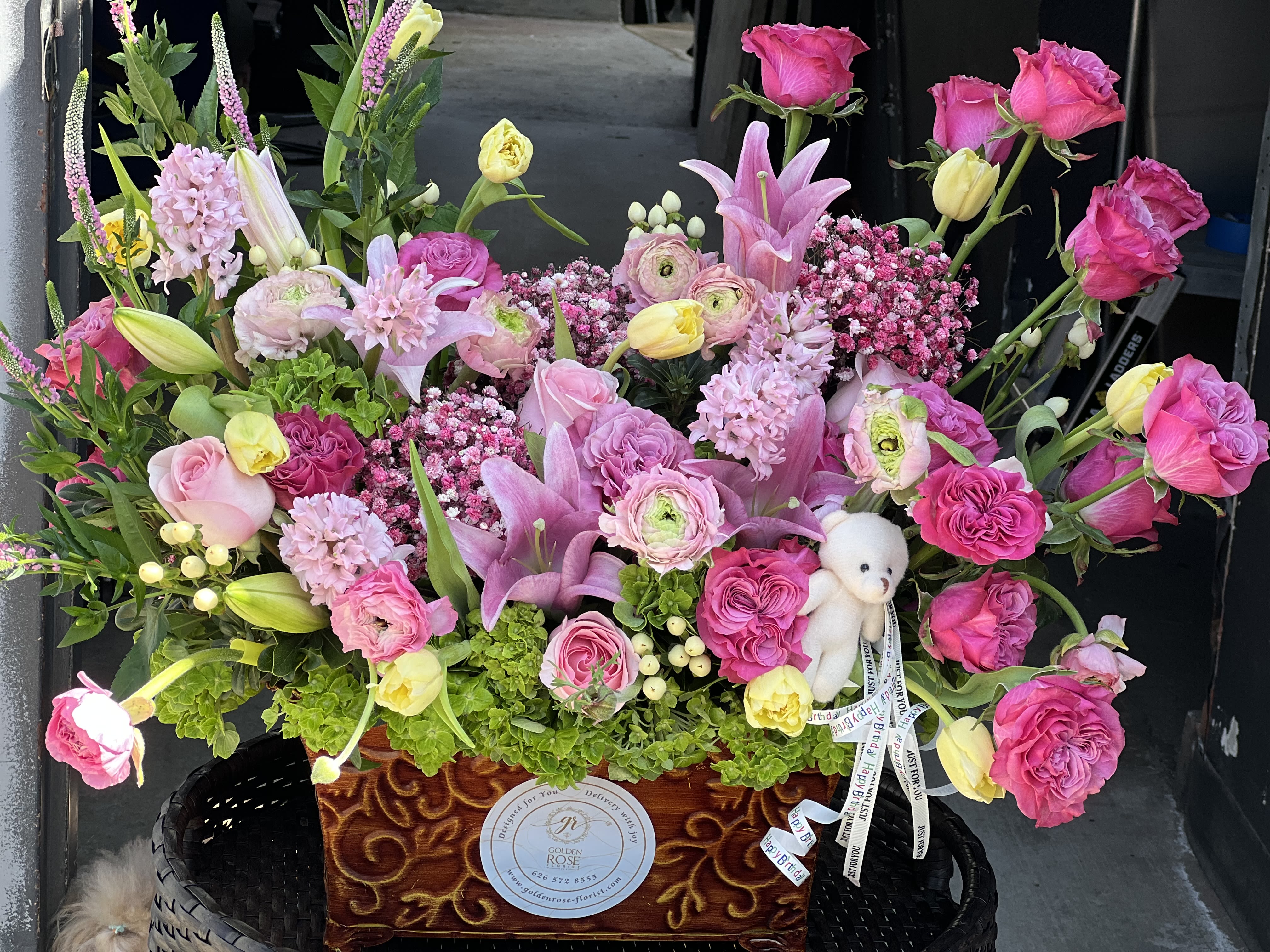 Blushing Bloom Basket - Soft, elegant, and overflowing with charm — this lush arrangement is designed to celebrate life’s sweetest moments. Featuring a radiant mix of pink roses, lilies, ranunculus, tulips, hyacinths, and seasonal textures, it’s lovingly arranged in a decorative wooden box and accented with a cuddly plush bear for an extra touch of sweetness.  Perfect for birthdays, baby showers, or simply to say “you’re loved.”   Arrangement includes: 	•	Premium roses, lilies, tulips, ranunculus,  	•	Lush seasonal greenery 	•	Keepsake wooden box 	•	Small plush bear (style may vary)  Substitutions may be made based on seasonal or in-store availability. The arrangement will be delivered as similar as possible.