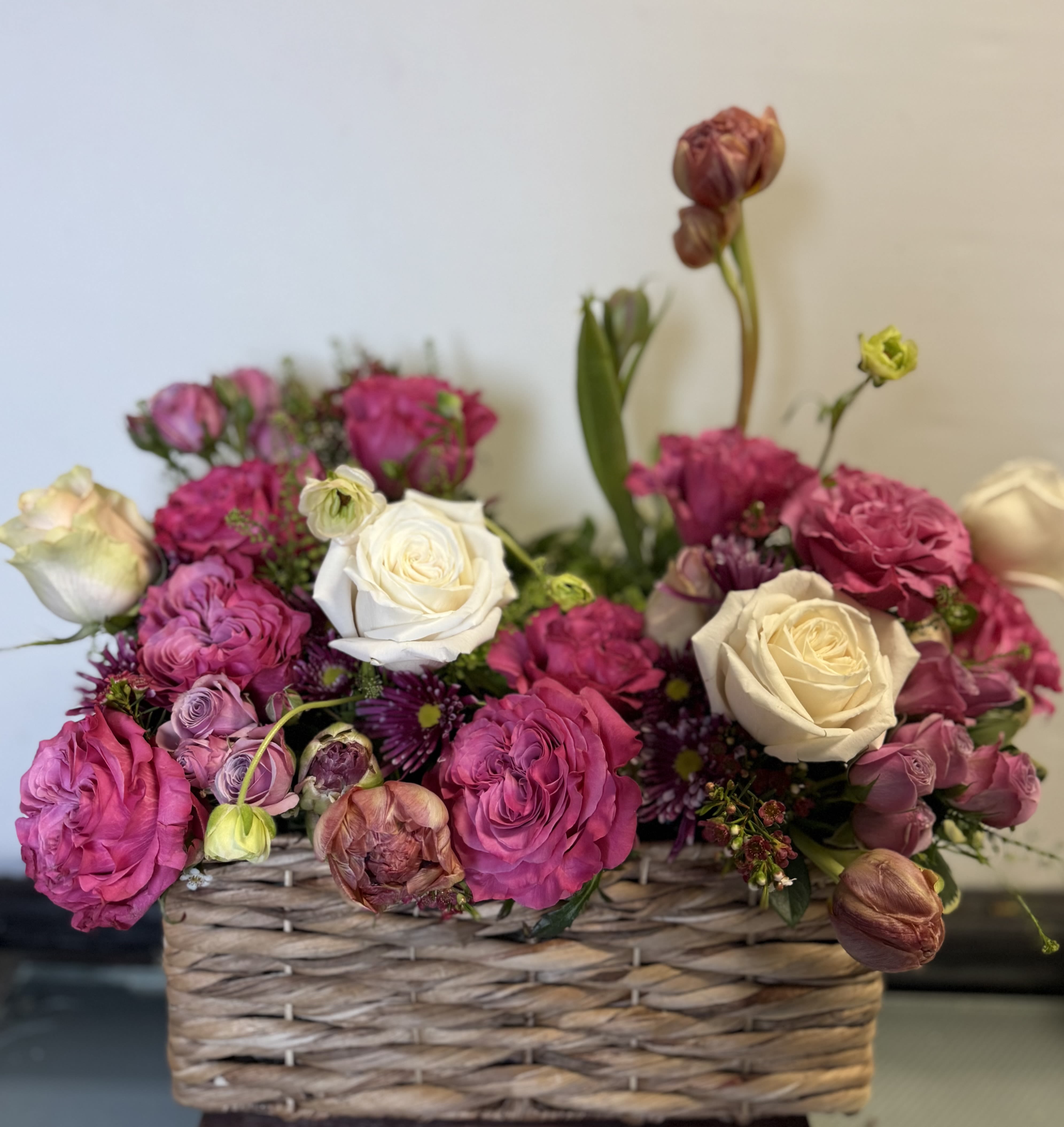 Blushing Elegance Basket - Bring warmth and romantic charm into any space with our Blushing Elegance Basket. This premium floral arrangement features a stunning mix of creamy white roses, deep magenta garden roses, lavender ranunculus, and seasonal tulips nestled in a rustic woven basket. Perfect for celebrating love, birthdays, or simply brightening someone’s day with a burst of elegance and color. A thoughtful and luxurious gift that speaks volumes without saying a word.  Substitution Note: Substitutions may be made based on seasonal or in-store availability. The arrangement will be delivered as similar as possible