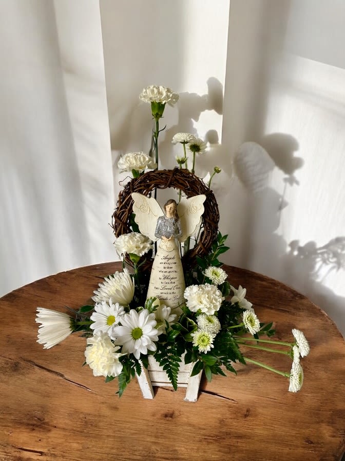 Heavenly Angel Memorial Arrangement - Heavenly Angel Memorial Arrangement  A gentle tribute for a cherished soul  Honor a loved one’s memory with our Heavenly Angel Memorial Arrangement, a peaceful and elegant display designed to bring comfort and serenity. Nestled in a rustic white wood container, this heartfelt design features a keepsake angel figurine surrounded by a delicate halo of natural grapevine, pure white carnations, daisies, mums, and soft greenery.  At the heart of the design stands a beautifully crafted angel inscribed with an uplifting message of remembrance—perfect for celebrating a life that continues to inspire.   Features:    •   Fresh white blooms for purity and peace    •   Inspirational angel statue keepsake    •   Gracefully arranged with care and compassion    •   Ideal for memorial services or as a sympathy gift  Whether placed at a service, or gifted to a grieving family, this arrangement speaks softly but profoundly—offering a message of love, hope, and eternal light.  ️ “Angels are always near to those who are grieving, to whisper to them that their loved ones are safe in the hands of God” is the sweet and comforting message inscribed on the angel. 