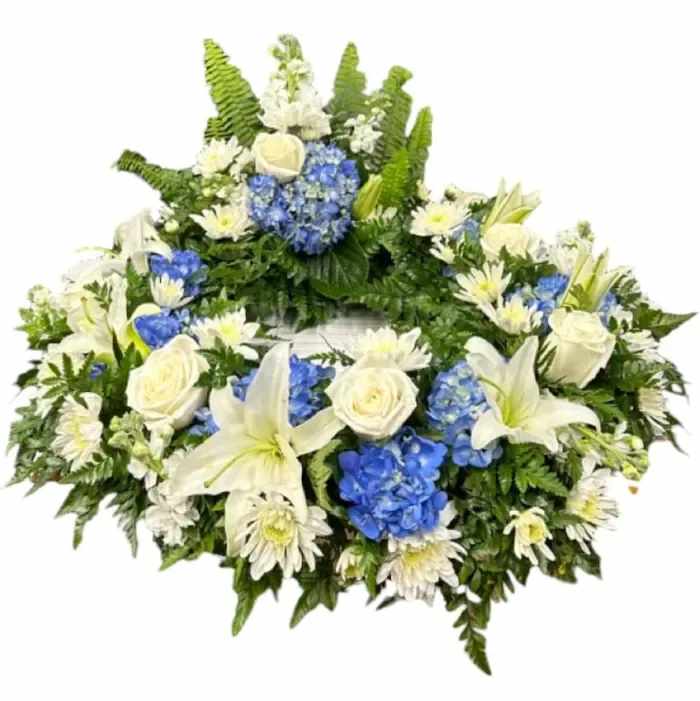 Honored Legacy Urn Wreath - The &quot;Honored Legacy&quot; urn wreath features a graceful combination of white lilies, blue hydrangeas, white mums, and verdant greenery, symbolizing purity, gratitude, respect, and renewal, respectively. With delicate lilies as its centerpiece, surrounded by the serene blue of hydrangeas and the purity of white mums, the arrangement offers solace and reverence to those mourning the loss of a loved one. The inclusion of lush greenery serves as a reminder of the enduring cycle of life. Together, these elements create a poignant tribute, honoring the legacy of the departed and providing comfort to grieving hearts.