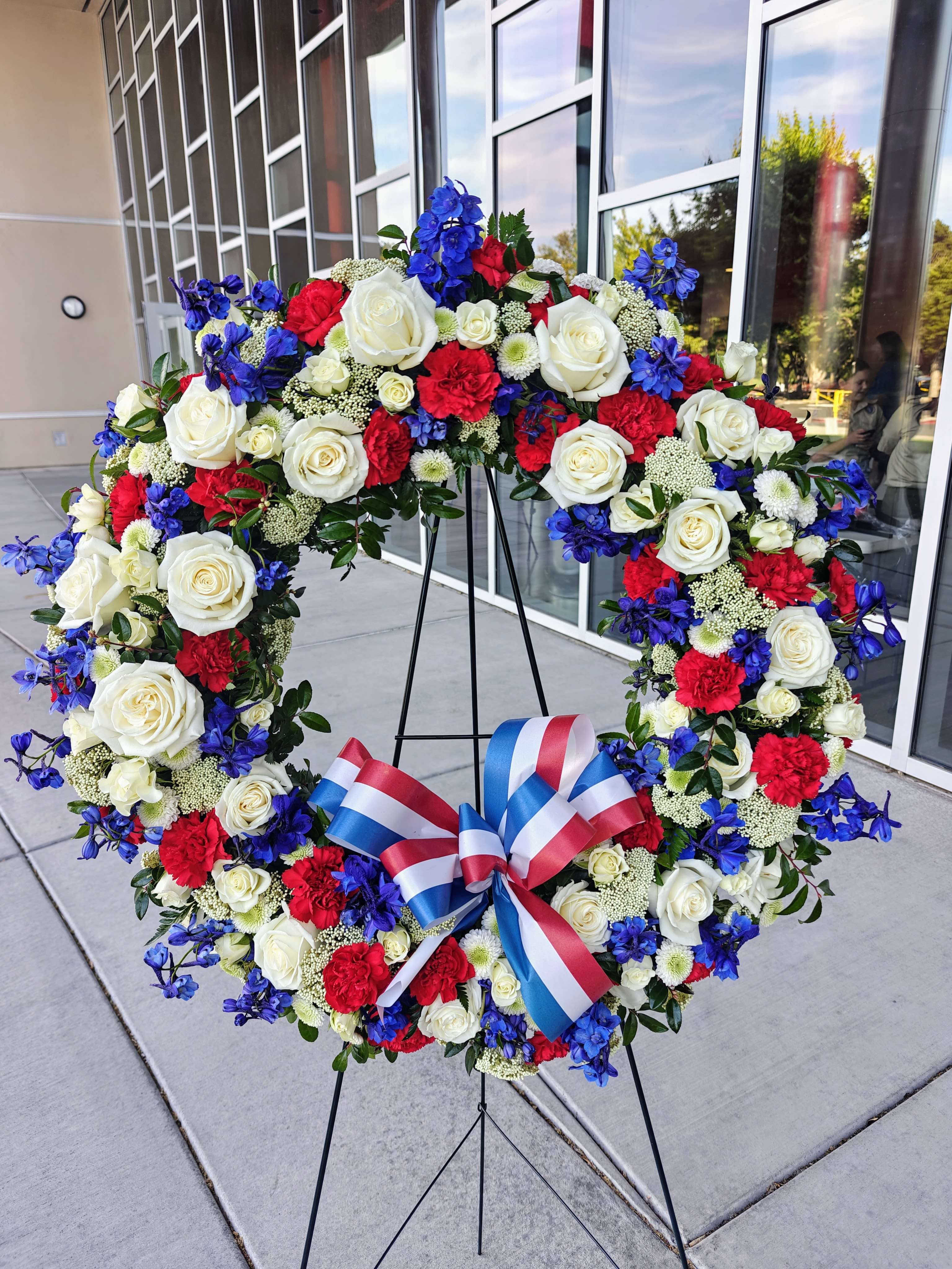 Large Red White and Blue Memorial Wreath - Memorial Wreath, approximately 25” wide. Stands on a tall Easel. Red White and Blue patriotic display of Flowers and bow. Be sure to order well in advance as blue flowers are hard to come by.