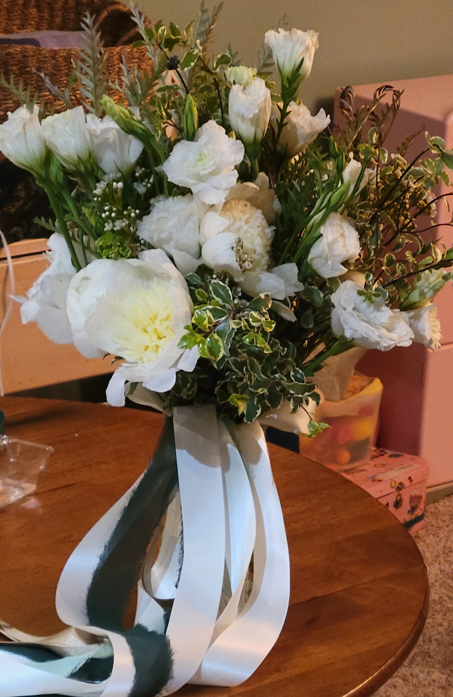 Bridal Hand-tied Bouquet - Soft, romantic, and timeless—this hand-tied bouquet showcases the classic beauty of lush white peonies and delicate lisianthus, accented with sprigs of greenery and variegated foliage. The full blooms are perfectly complemented by airy textures and a luxurious cascade of satin ribbons in soft white and deep green tones.  