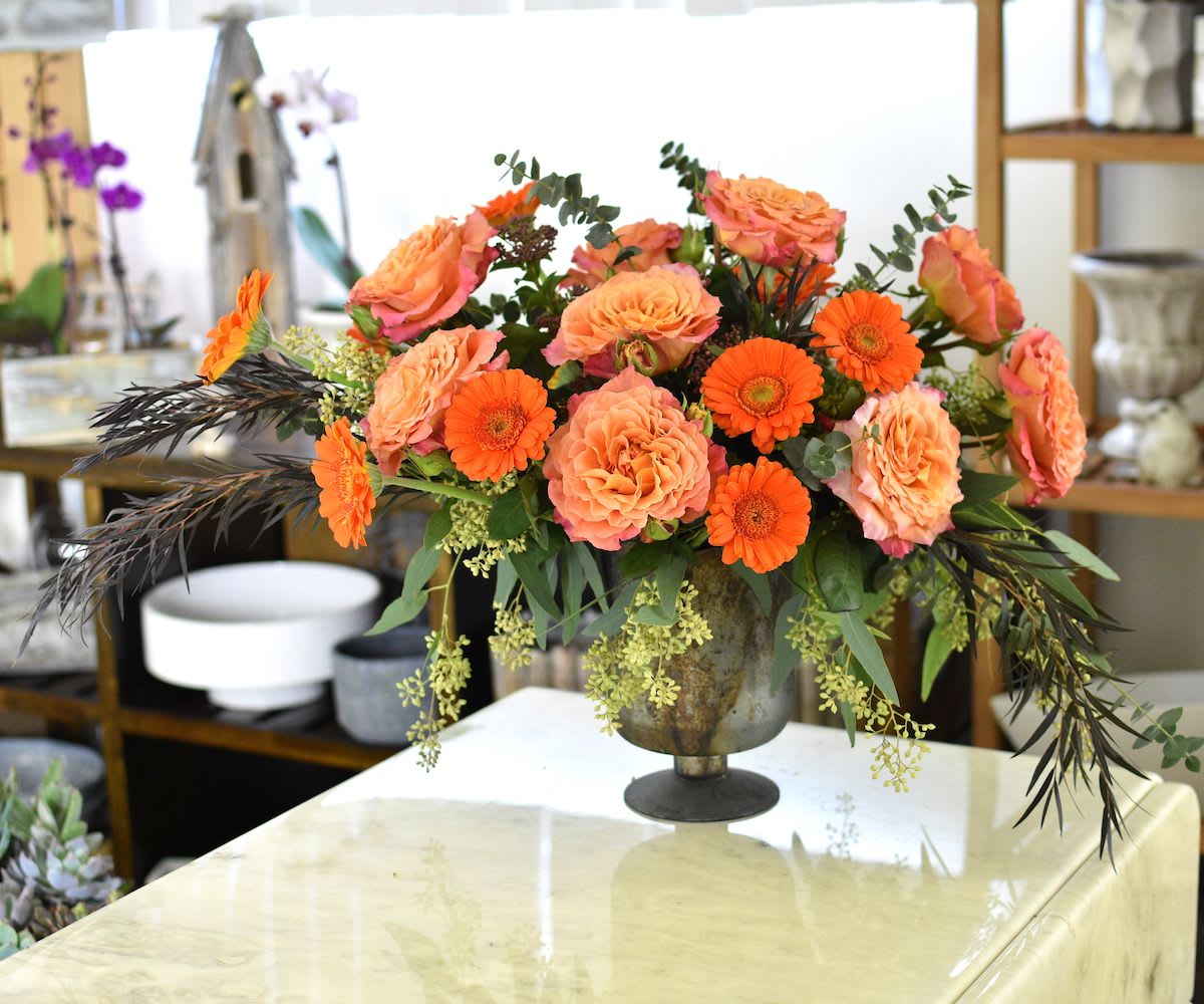 Chelsea 1 Chelsea 1 - Radiant Ember in Antiqued Chelsea Vase A bold and elegant display. Showcases vibrant orange gerberas and lush roses in warm coral tones, accented by dramatic dark foliage, eucalyptus, and delicate seasonal textures. Set in an antiqued Chelsea vase, this arrangement strikes a perfect balance between timeless charm and modern sophistication. Ideal for making a lasting impression—whether for a celebration or simply to brighten a space with refined beauty.