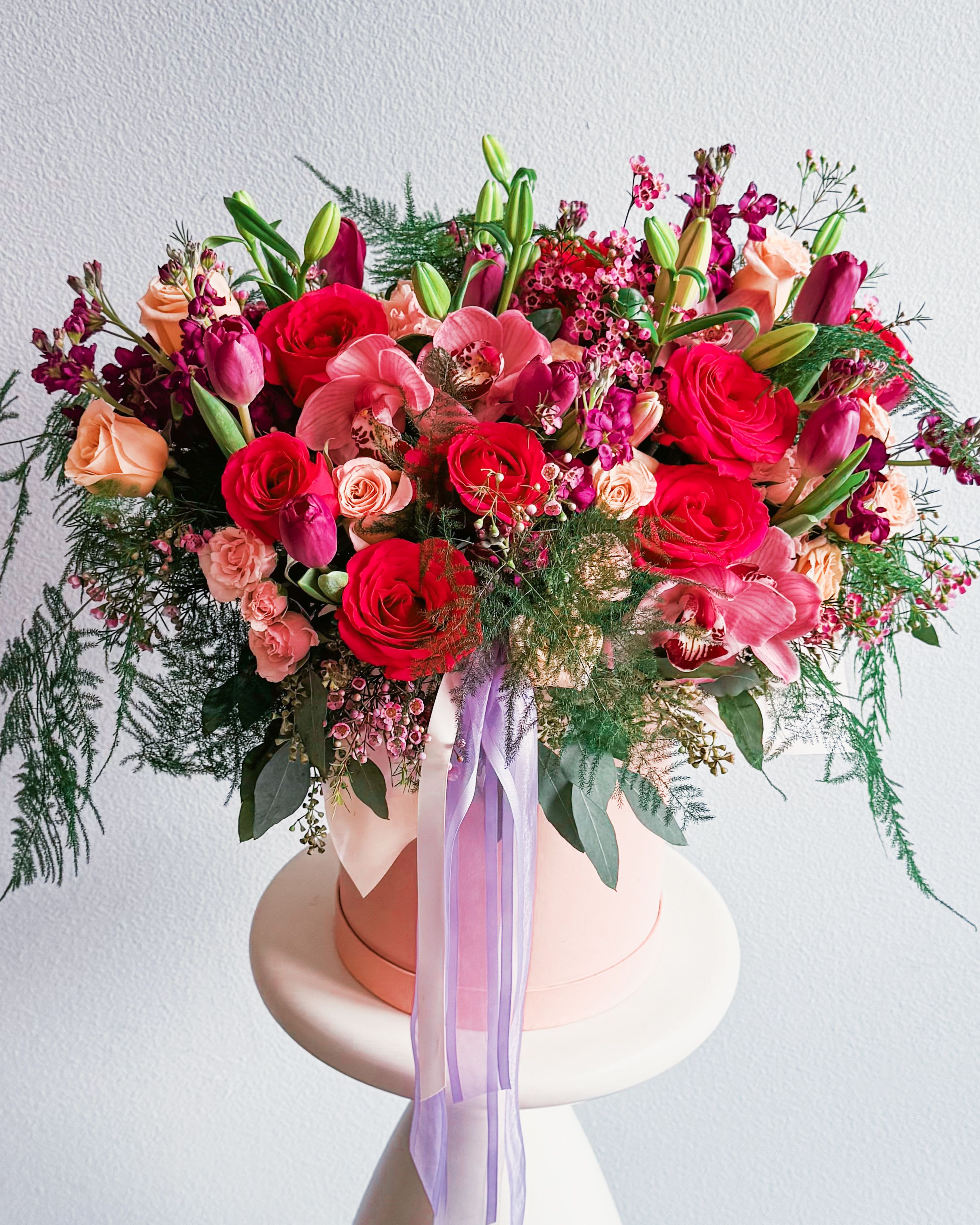 Vivid Velvet - This bouquet is a total showstopper! With its fiery red roses, soft peach spray roses, and bold magenta tulips, it’s got that &quot;can’t take your eyes off it&quot; kinda vibe. The lush orchids and delicate waxflowers bring in a fancy touch, while the wild greenery keeps it fresh and fun. Wrapped up in a chic pastel hatbox with a flowing lilac ribbon, this beauty is pure floral magic.