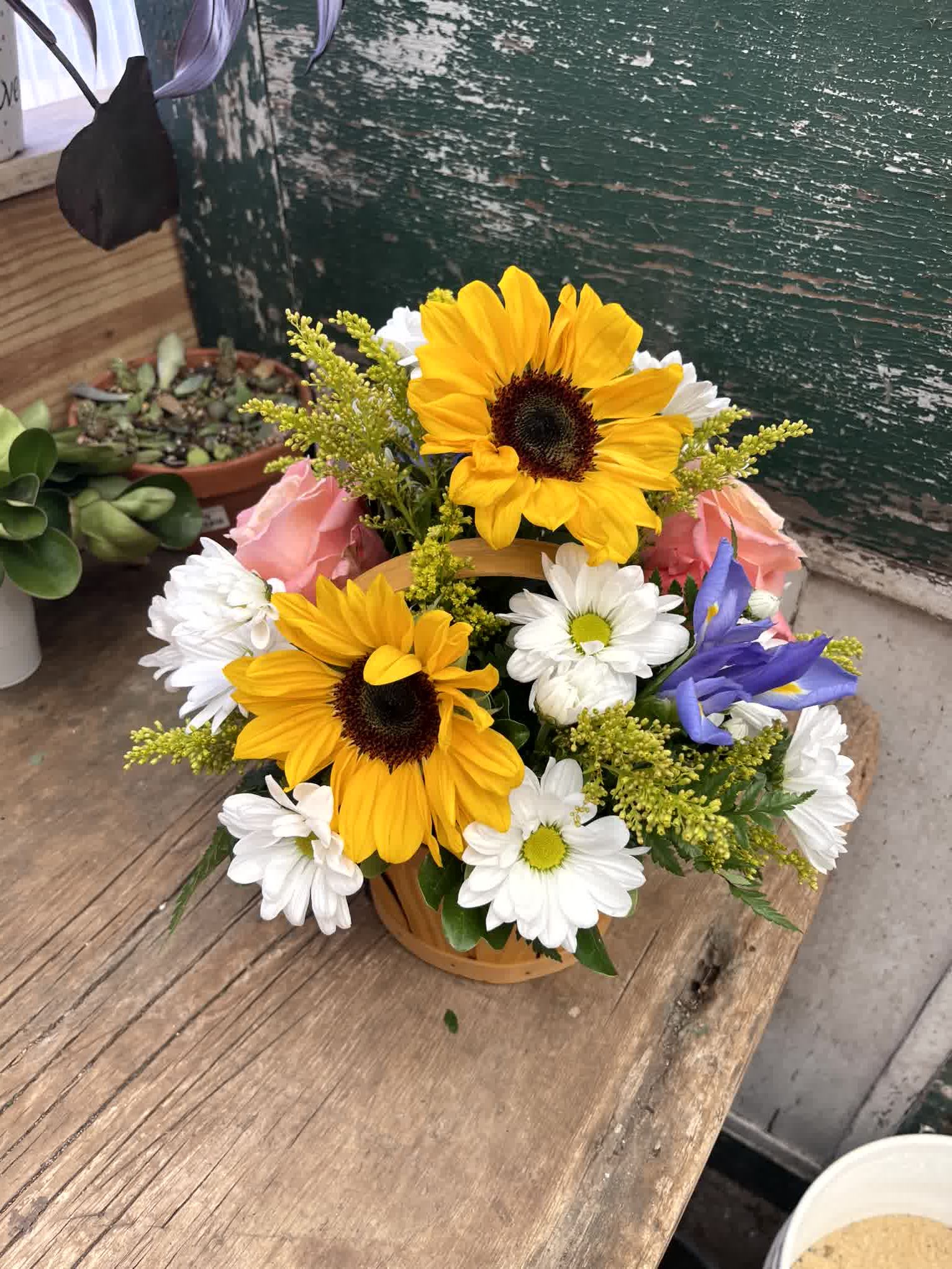Birthday Blast - A bright, happy basket arrangement that will warm the heart of the sunshine in your life. Beautiful sunflowers, happy daisies, blue iris, roses, solidago and greenery are sure to make her smile on her special day. 