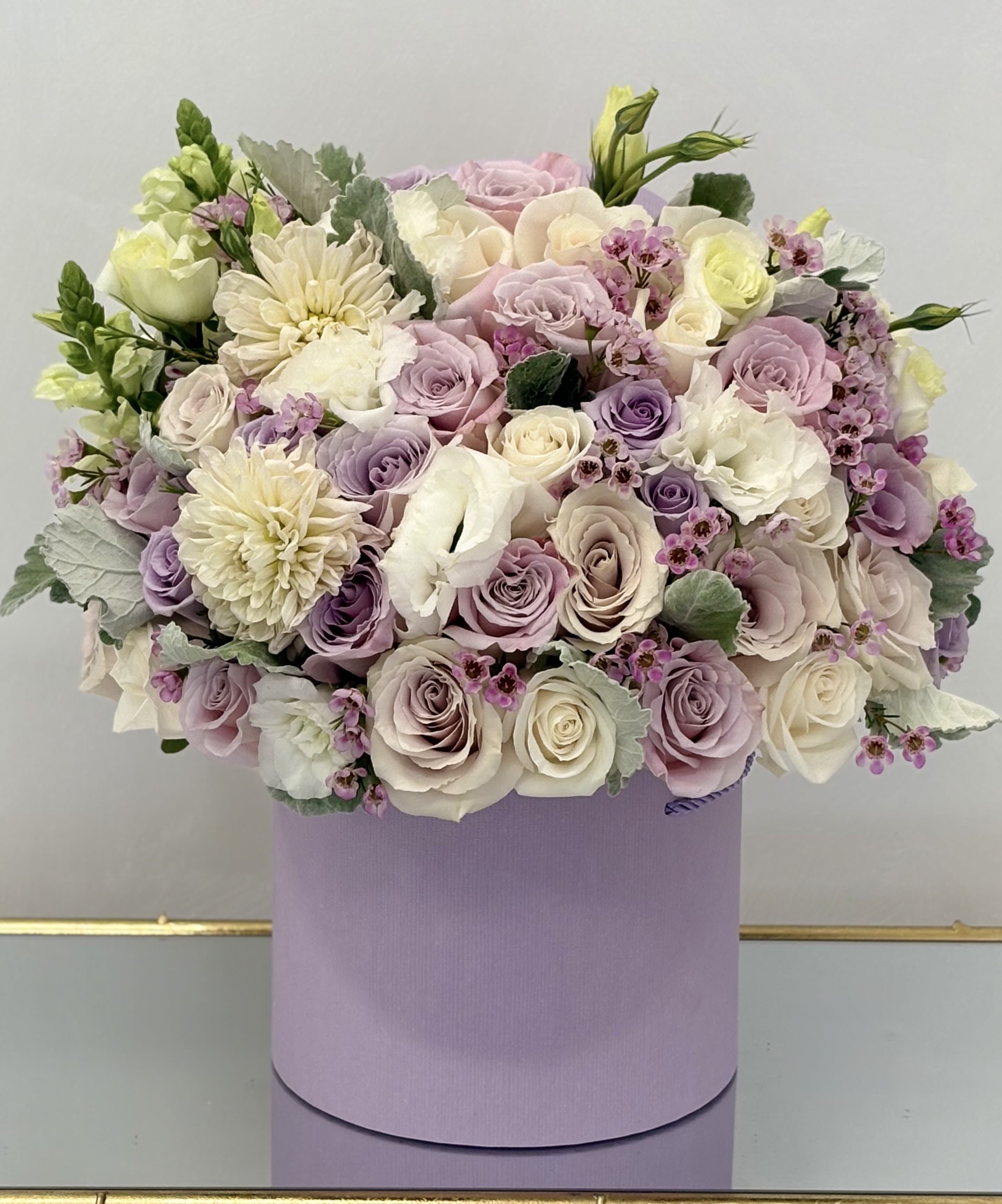Lavender Whispers Hat Box By VLM Flowers - A graceful medley of soft lavender, blush, and cream blooms, Lavender Whispers is a floral daydream in a lilac hat box. Featuring lush roses, creamy dahlias, lisianthus, and waxflowers, this arrangement is a celebration of calm beauty and refined charm. Accents of dusty miller add a silvery softness, making it a perfect gift for anniversaries, sympathy gestures, or simply saying “you’re loved.”