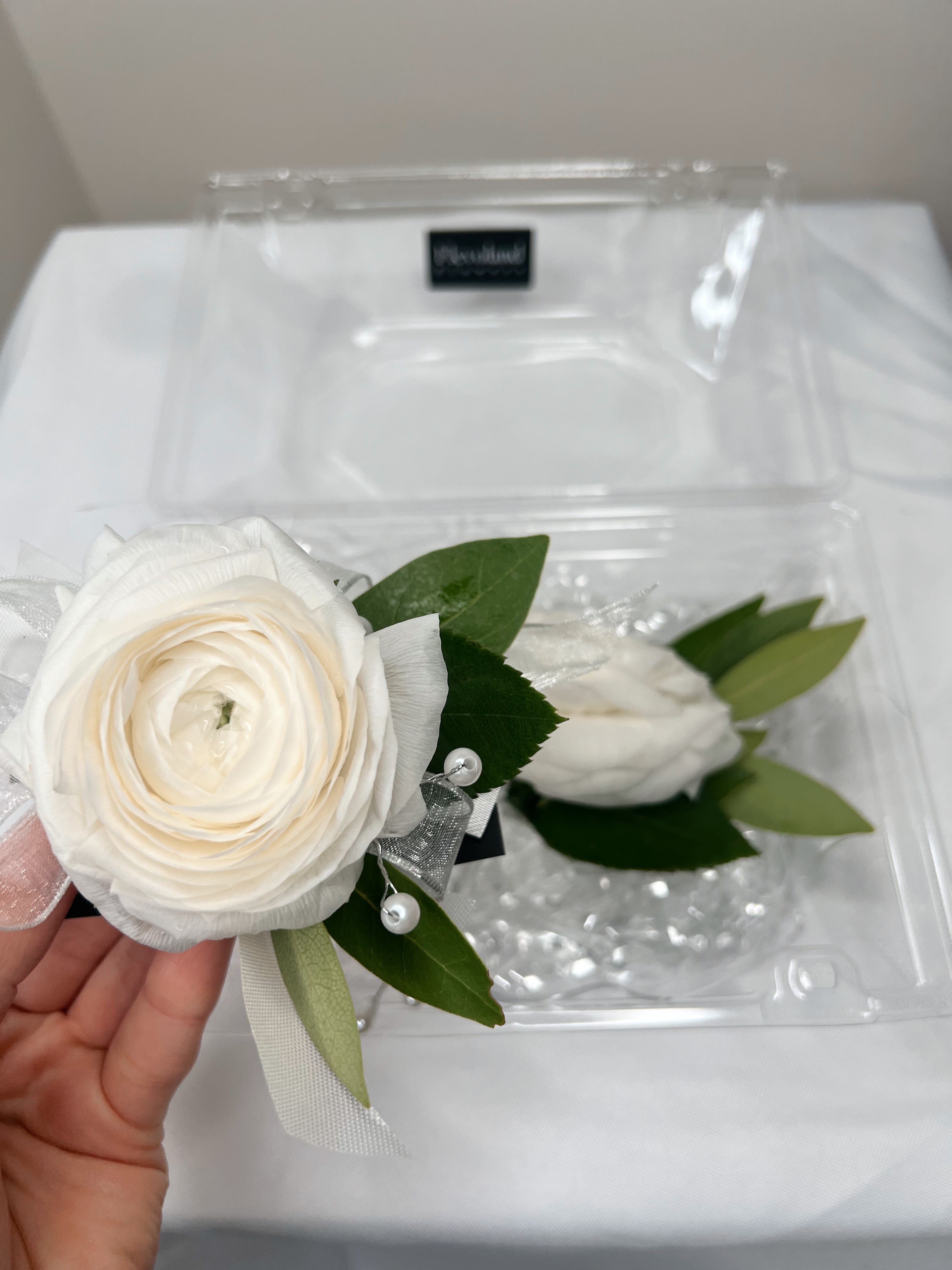 White Ranunculus Corsage and Boutonnière - This elegant white ranunculus corsage and boutonnière are the perfect choice to add a touch of sophistication to any outfit. The ranunculus, with its delicate beauty, paired with a clean and elegant design, creates a harmonious set ideal for special occasions like formal events.