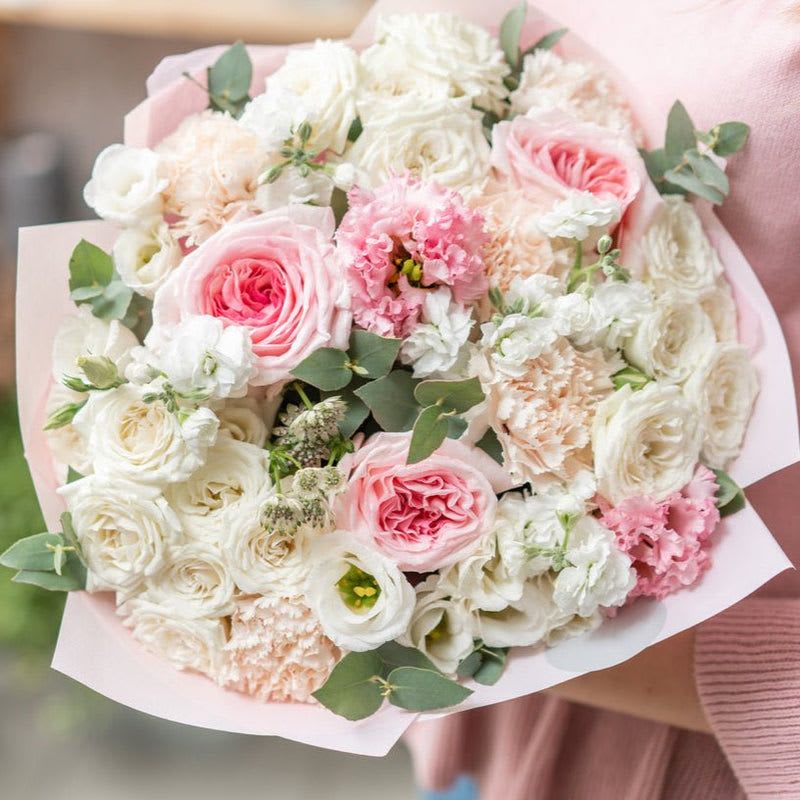 Essence of Elegance - Introducing a stunning hand-tied bouquet that captures the essence of elegance and romance through its delicate composition. This exquisite arrangement features pristine white roses, complemented by soft lisianthus, light pink carnations, and enchanting garden roses in shades of pink. The combination of these blooms creates a harmonious blend that conveys beauty and charm.  The white roses symbolize purity and new beginnings, serving as a perfect foundation for this thoughtful bouquet. Paired with the airy lisianthus, known for its graceful, cup-shaped blossoms, the arrangement gains a touch of sophistication. The light pink carnations add a playful softness, representing a deep admiration and a sense of joy, while the lush garden roses in varying pink hues infuse the bouquet with romance and depth.  Lovingly hand-tied and wrapped in natural paper, this bouquet emanates rustic charm while showcasing the meticulous care taken in its construction. The gentle hues and lush textures create a visual feast that is both captivating and heartwarming. Whether marking a special occasion, expressing gratitude, or simply brightening someone’s day, this hand-tied bouquet serves as a beautiful expression of love and appreciation, certain to bring joy to anyone fortunate enough to receive it.