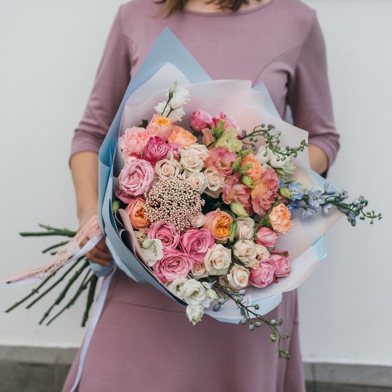 Celebration of Love - This hand-tied bouquet is a romantic arrangement that exudes elegance and charm, perfect for any special occasion. At the heart of the bouquet, lush roses in varying shades of creamy pink and soft peach create a lovely focal point, their velvety petals layered gracefully. Accompanying these stunning blooms are delicate garden roses, which add a vintage flair with their ruffled petals and charming fragrance, further enhancing the ethereal quality of the bouquet.  Interspersed among the roses, the graceful spikes of delphinium introduce a touch of height and texture, their soft blue hues complementing the warm tones of the pink and peach roses beautifully. The airy presence of rice flower lends a whimsical touch, its tiny white blossoms creating a gentle, cloud-like effect that adds lightness to the arrangement.  Wrapped gently with natural twine or a soft chiffon ribbon, the bouquet captures the essence of a dreamy garden, with its harmonious blend of colors and textures creating a heartwarming and inviting aesthetic. This exquisite arrangement is sure to enchant anyone who beholds it, making it a perfect gift or centerpiece for weddings, showers, or any celebration of love.