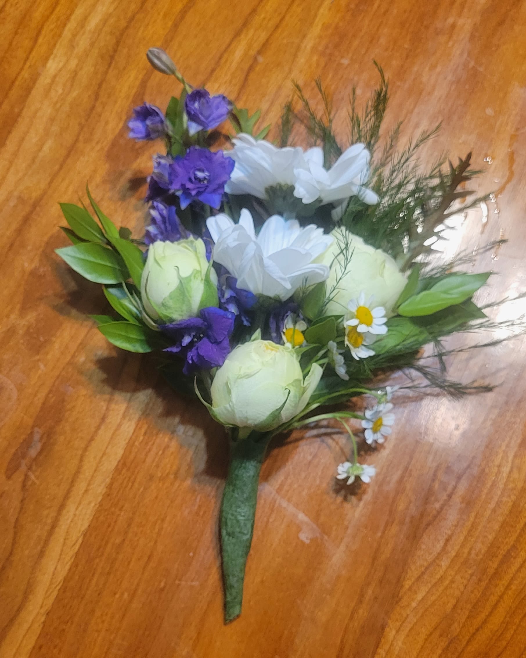 Mother's Delight by Dixie's Flowers - Delicate and full of charm, this hand-tied corsage captures the fresh beauty of a spring garden. It features creamy white rosebuds, cheerful white daisies, vibrant purple larkspur, and soft greenery, all carefully arranged for a natural, elegant look.  