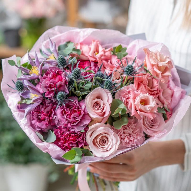 Blushing Elegance - This arrangement is a delightful symphony of pink hues, combining elegance and charm with a touch of modern flair. At its core, pink roses in varying shades create a lush and romantic focal point, their layered petals radiating timeless beauty. Surrounding them, carnations in a spectrum of pink tones—from soft blush to vibrant magenta—add texture and depth, infusing the bouquet with warmth and vibrancy.  Pink lisianthus, with its delicate and flowing petals, introduces a sense of softness and refinement, enhancing the romantic feel of the design. To add a striking contrast, blue eryngium brings a unique, spiky texture and a cool tone, creating a contemporary balance to the arrangement's warm palette.  Perfect for any occasion, this bouquet captures the essence of elegance with a playful twist. Whether for celebrations, heartfelt gestures, or simply to brighten someone’s day, its harmonious mix of colors and textures makes it truly unforgettable.