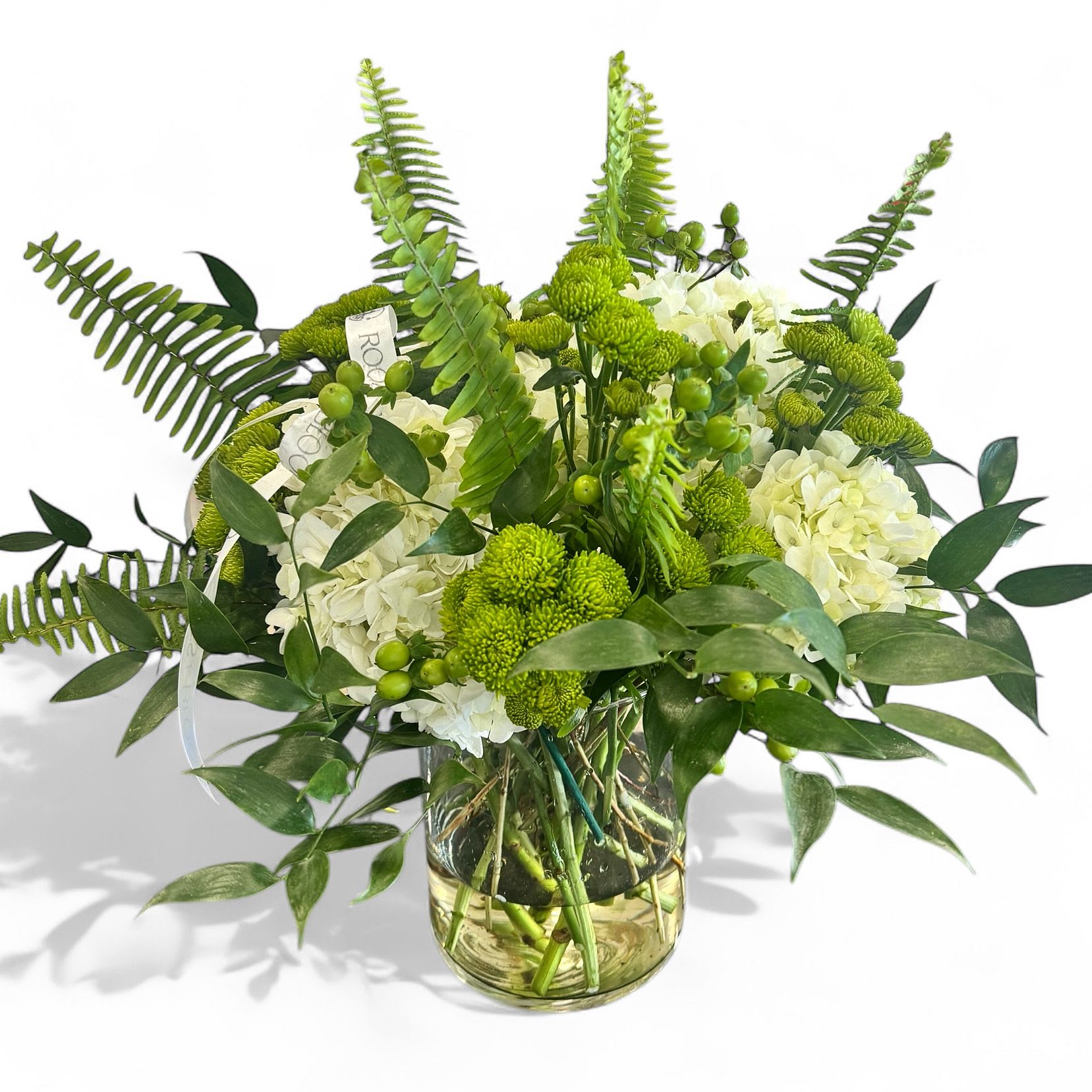 Green Muse - This arrangement is fresh, wild, and full of life! With its mix of white hydrangeas, green button chrysanthemums, berries, ferns, and assorted lush greenery in a clear glass vase, it feels like a serene walk through the forest clearing. 