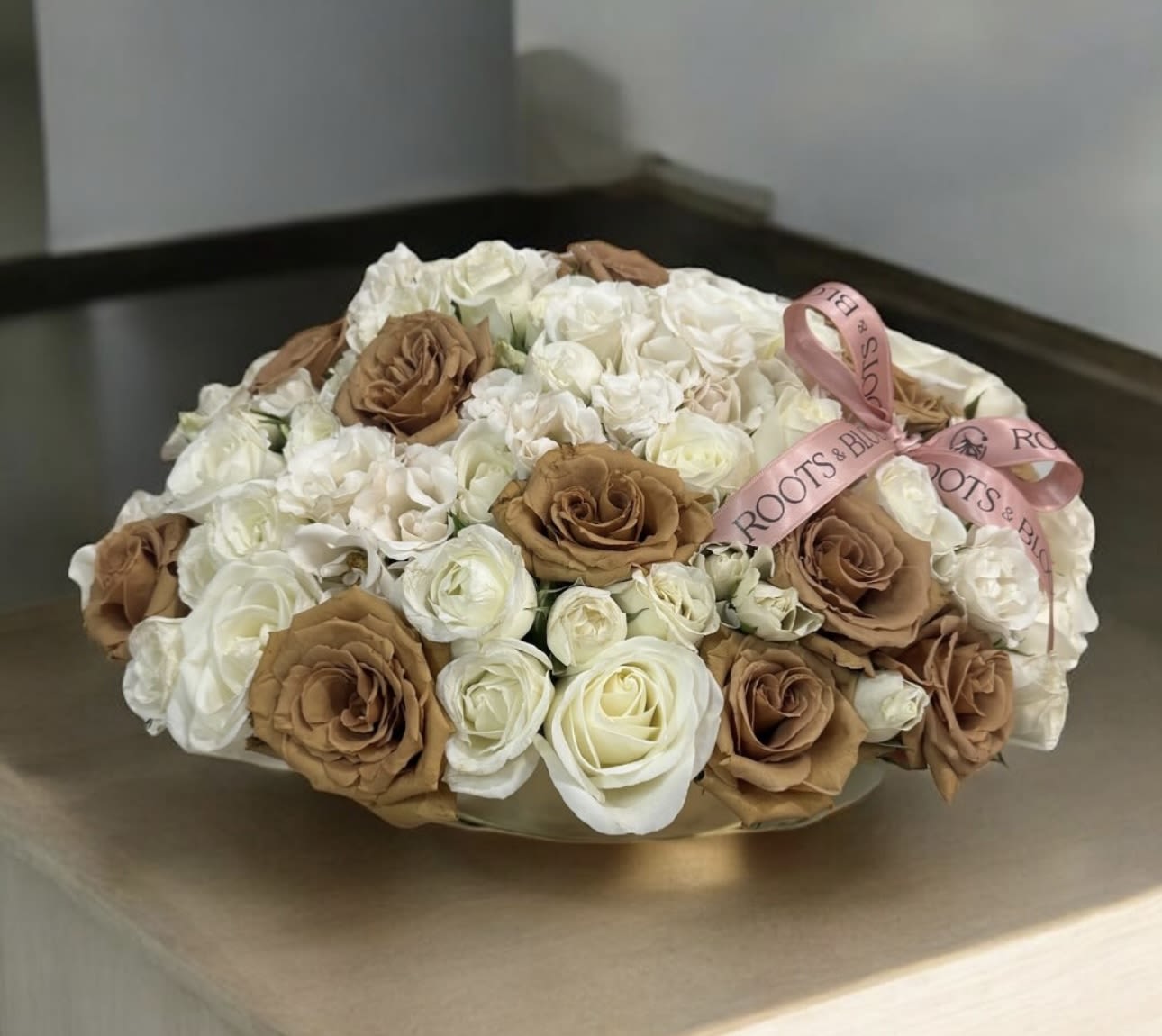 Golden Hour - Inspired by the warm glow just before sunset, Golden Hour brings timeless beauty into focus. Featuring creamy, white roses, delicate baby white blooms, and rich toffee-colored roses, this arrangement rests in a sleek, wide gold oval vase. It’s the perfect blend of elegance and warmth. -a statement piece radiates quite luxury.
