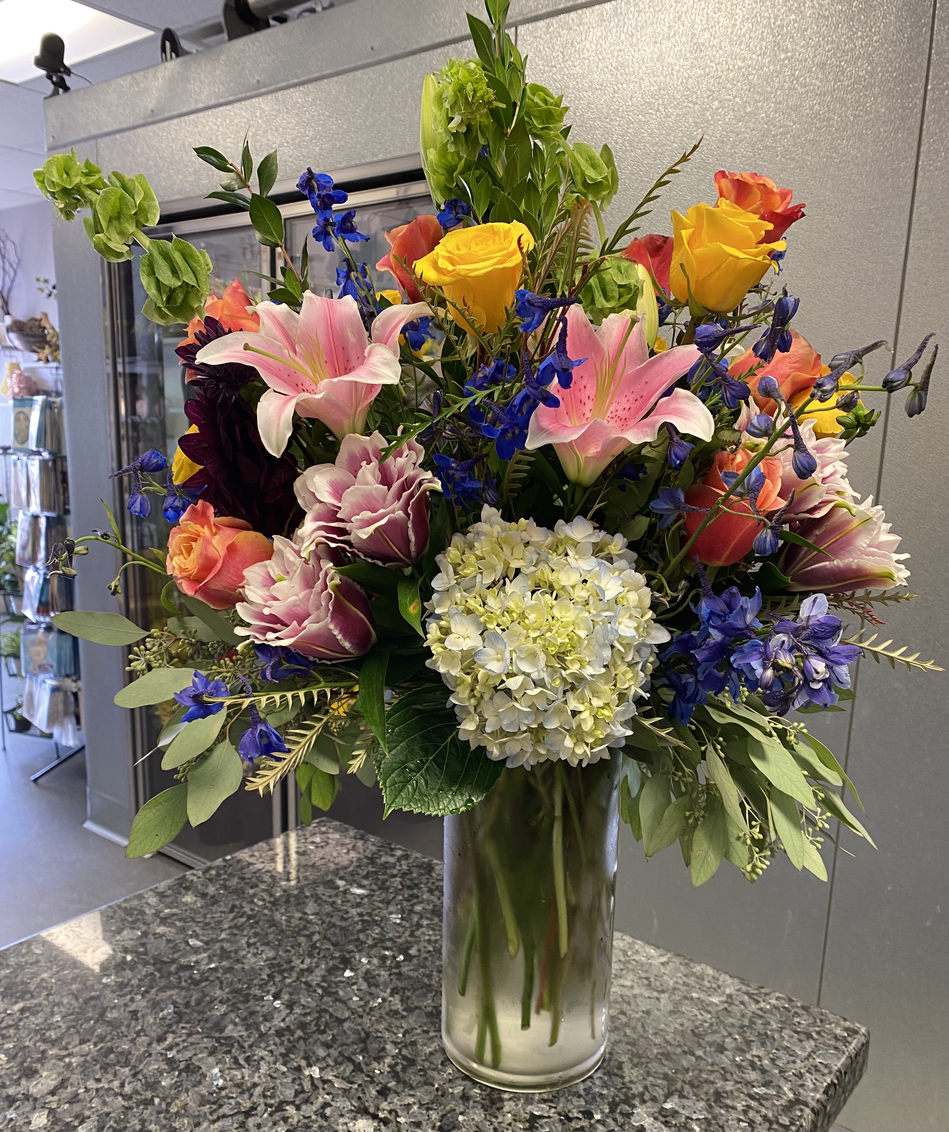 Bright and Fantastic - This Colorful Bouquet with Premium Flowers in a Tall Glass Vase includes Pink Lilies, Blue Hydrangea's, Bells of Ireland, Orange Roses, Yellow Roses, Blue Delphinium or as Similar as Possible.