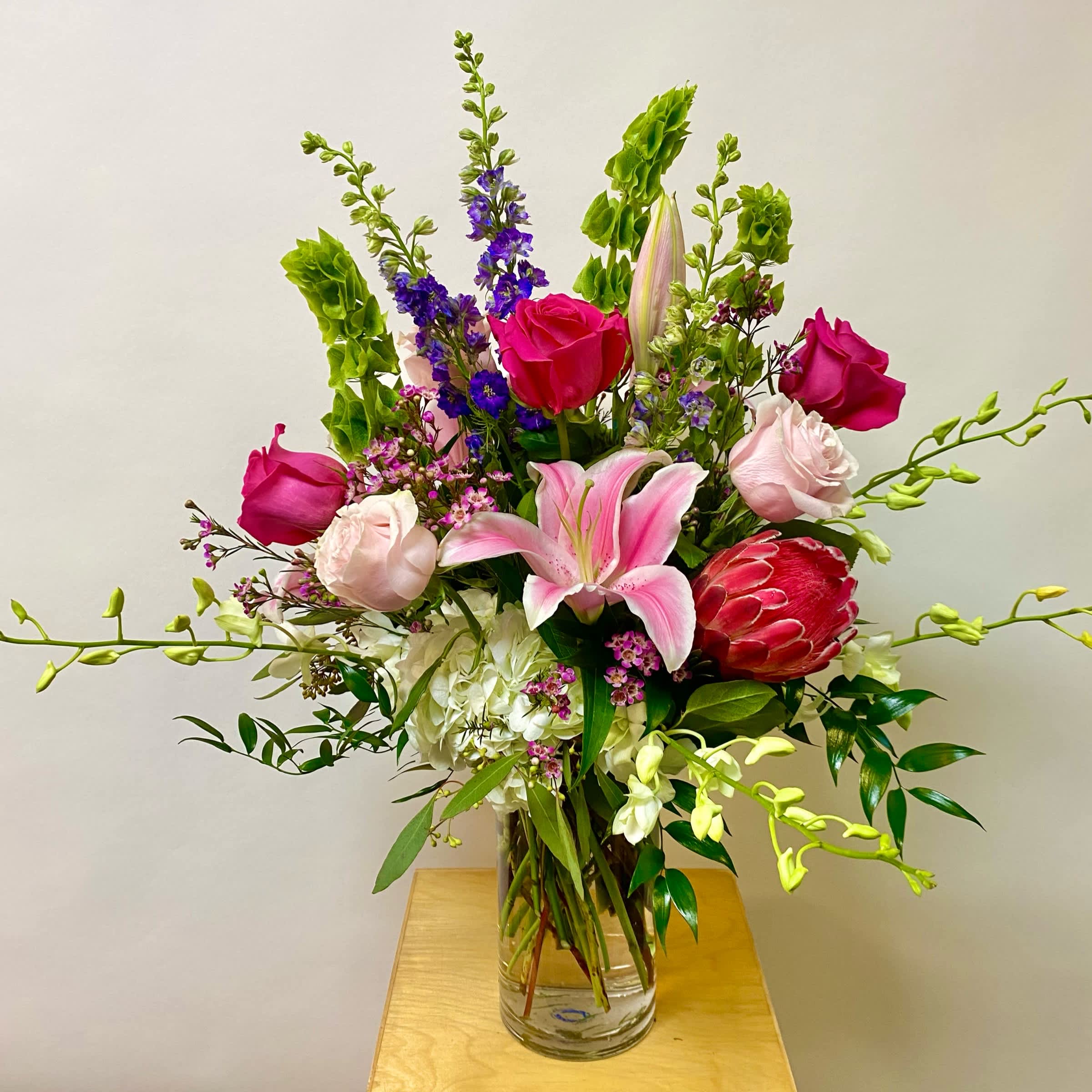 Springtime Embrace - This Springtime Arrangement Includes Protea, White Dendrobium Orchids, White Hydrangea's, Pink Lilies, Light Pink Roses, Hot Pink Roses, Purple Larkspur, Bells of Ireland, Wax Flower and Greenery in a Tall Glass Vase. Or as Similar as Possible. 