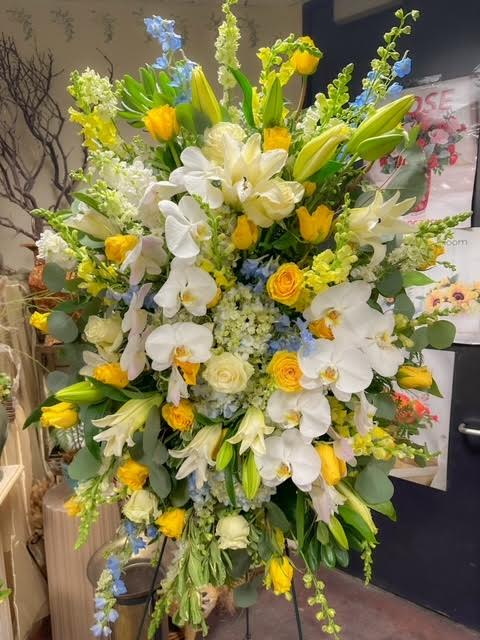 Serenity Bloom Standing Spray - The Serenity Bloom Standing Spray is a beautiful tribute to honor and celebrate a loved one's life. Crafted with an elegant blend of yellow roses, orchids, and white lilies, this arrangement brings a sense of peace and comfort to any memorial service. The soft, uplifting hues of yellow, blue, and white symbolize hope, serenity, and remembrance, creating a heartfelt expression of sympathy.