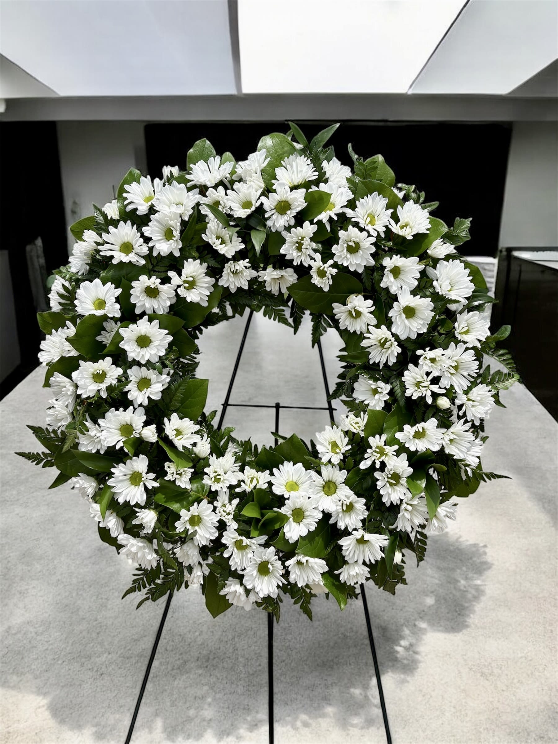 Daisy Garden Wreath/Jardin de Margaritas  - An elegant wreath crafted with fresh white daisies, symbolizing purity, peace, and everlasting love. The circular design represents the cycle of life and the continuity of remembrance. Accented with natural greenery, this wreath conveys serenity and respect, making it a beautiful tribute to honor and say farewell to a loved one.  Una elegante corona elaborada con delicadas margaritas blancas frescas, símbolo de pureza, paz y amor eterno. Su diseño circular representa el ciclo de la vida y la continuidad del recuerdo. Acompañada de follaje verde natural, esta corona transmite serenidad y respeto, convirtiéndose en un hermoso homenaje para despedir con dignidad a un ser querido.  