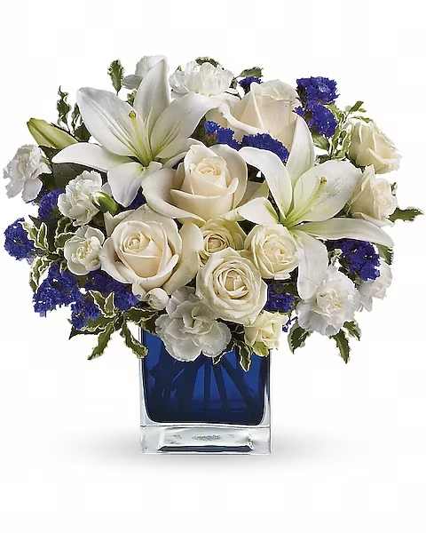 Teleflora's Sapphire Skies Bouquet - Send someone a bit of heaven with this beautiful bouquet. Luxurious crème roses and pure white lilies paint a peaceful picture inside a sapphire blue cube.  Crème roses, white asiatic lilies and white miniature carnations are mixed with bursts of purple statice and green pitta negra. Delivered in a glass Cube.