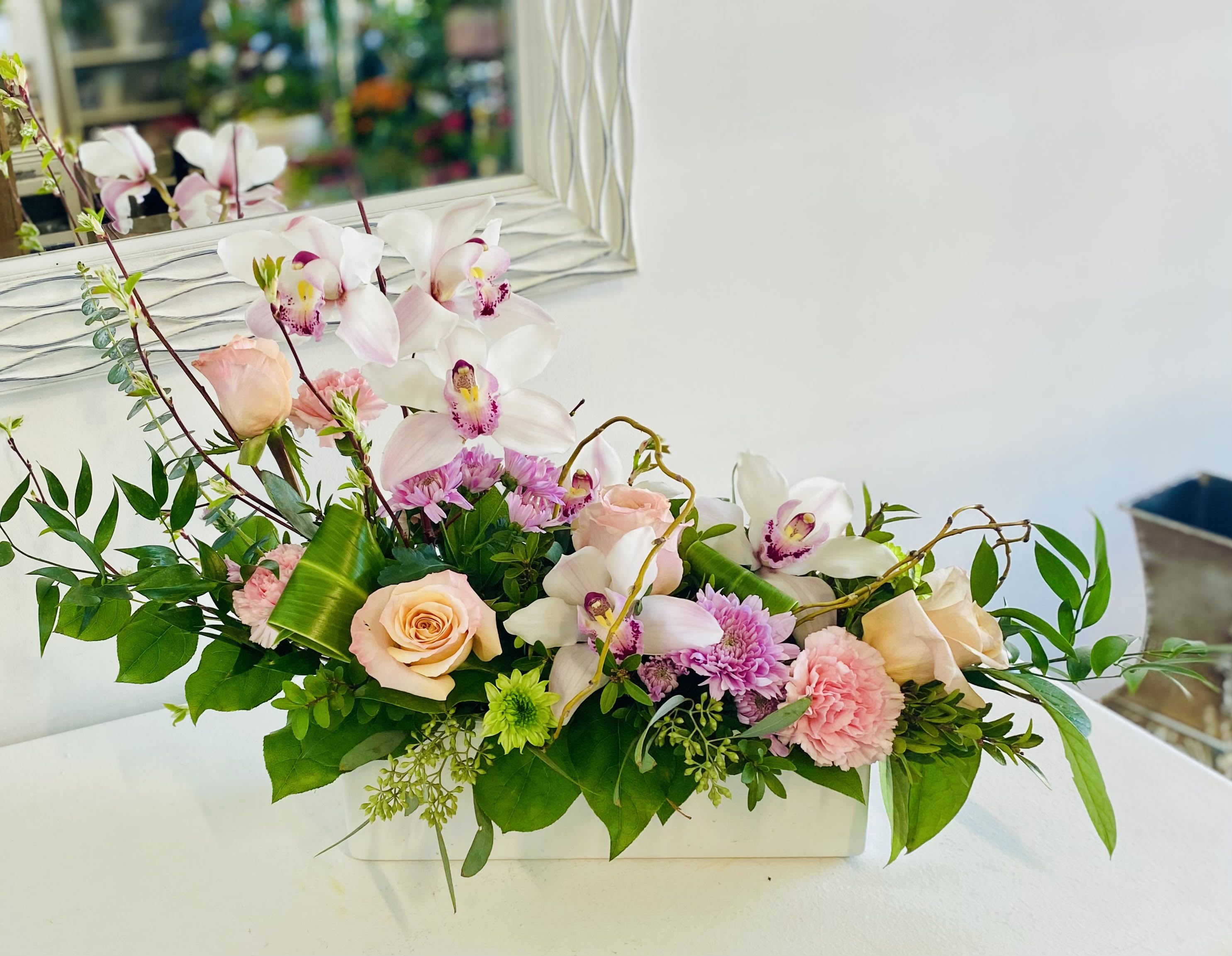 Peachy Blooms Peachy Blooms - This elegant and airy arrangement features graceful white and blush cymbidium orchids paired with soft peach roses, pink carnations, lavender mums, and delicate greenery. Accented with natural branches and displayed in a modern white ceramic container, it brings a touch of sophistication and tranquility—perfect for weddings, sympathy, or refined home décor.