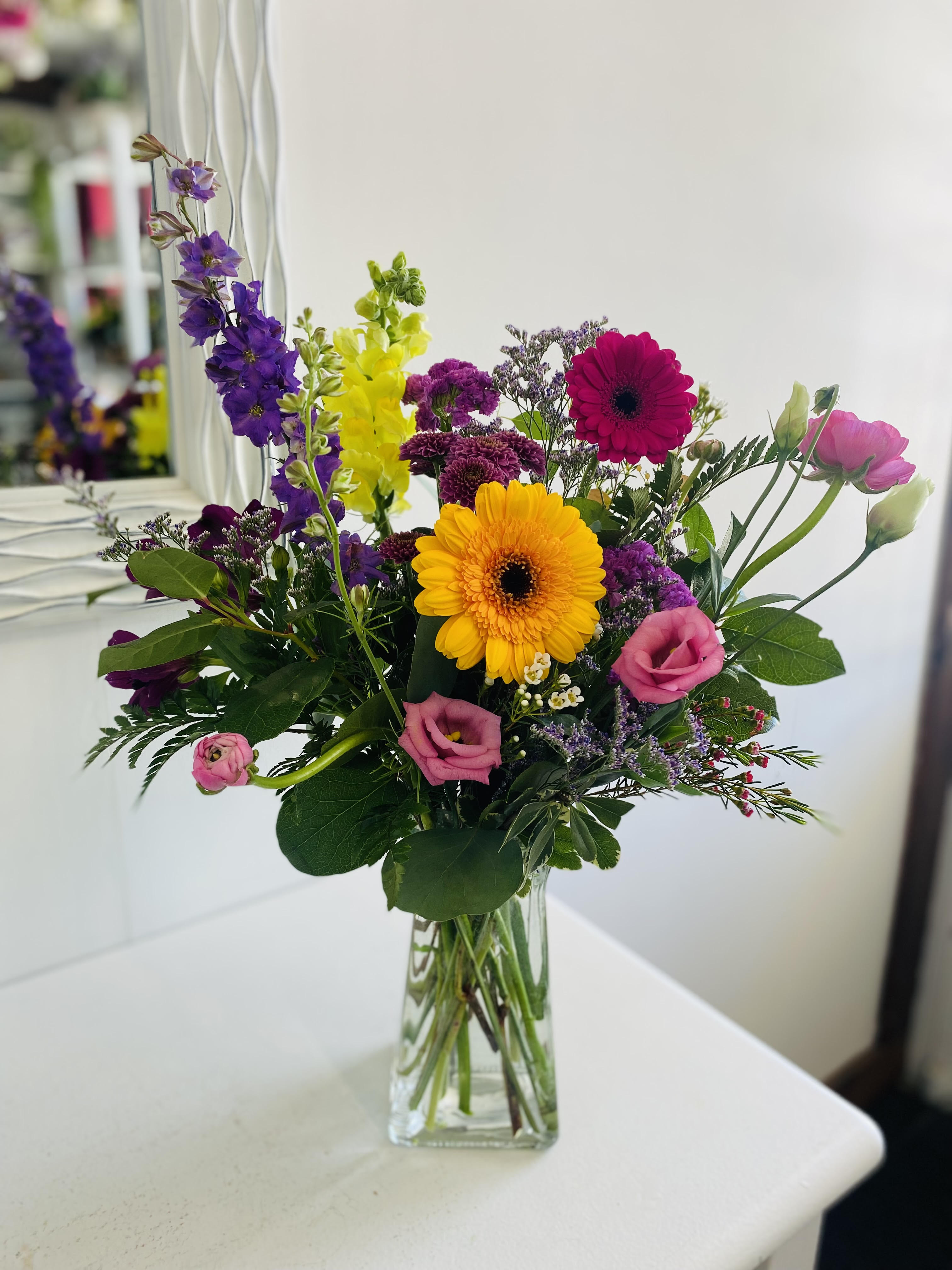 Wildflower Design Wildflower Design - This tall, cheerful arrangement is a burst of color and texture, featuring sunny yellow and magenta gerbera daisies, deep purple stock, bright yellow snapdragons, and soft pink lisianthus. Accented with delicate filler flowers and lush greenery, all elegantly arranged in a tall clear glass vase, it’s perfect for adding height and vibrance to any space or celebration.