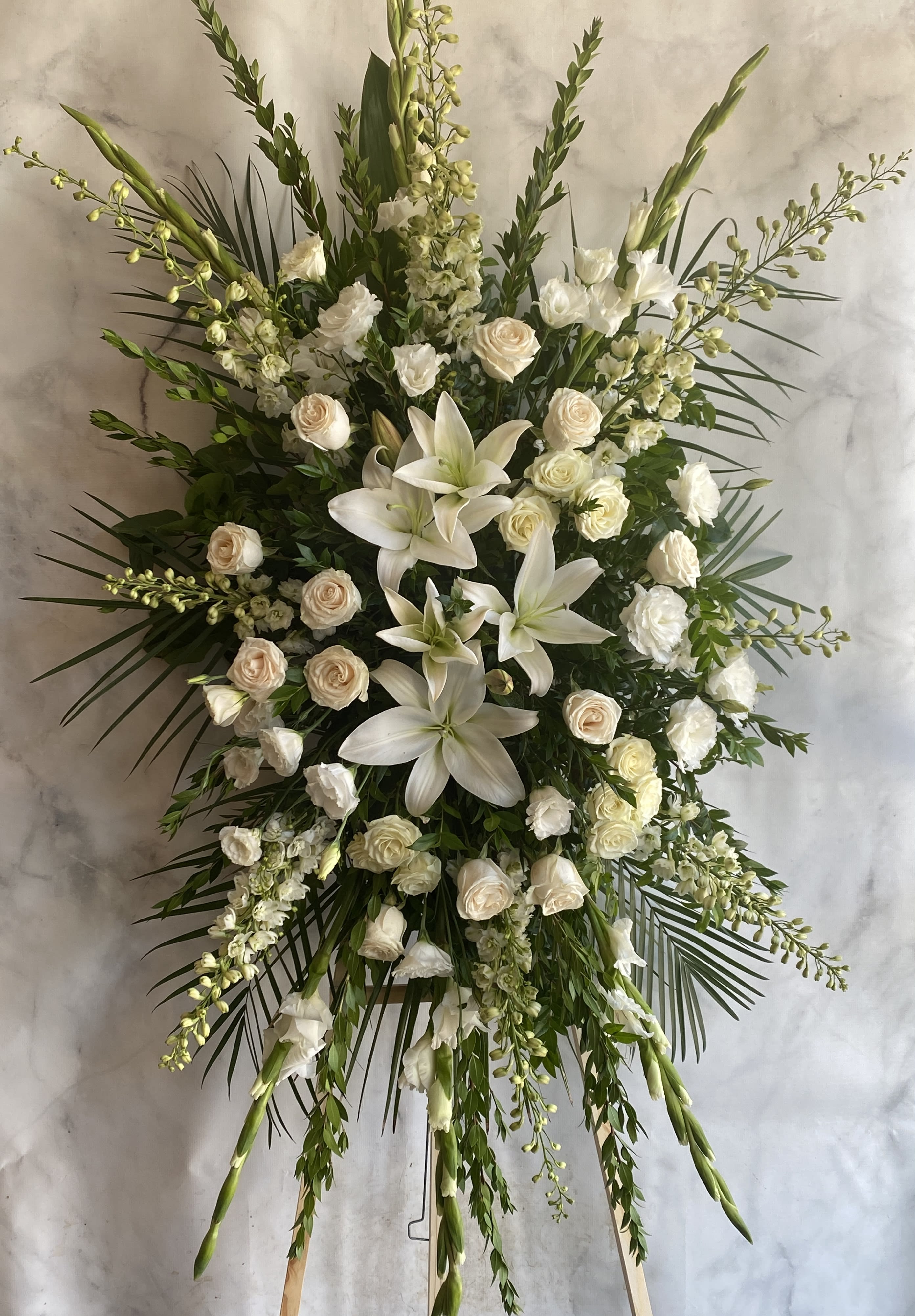 Loving Memory Spray - Experience the tranquility and purity of our white funeral arrangement. Delicate lilies, elegant roses, and charming mums come together to create a stunning display of love and respect. Please note that due to the natural variations in seasonal blooms, the flowers used in your arrangement may differ from those pictured on our website. Rest assured that we only use the freshest, most beautiful flowers available, and that your arrangement will be thoughtfully designed with the same color scheme and style. Standing approximately 4 feet tall and 3 feet wide, this arrangement will be a memorable tribute to your loved one.