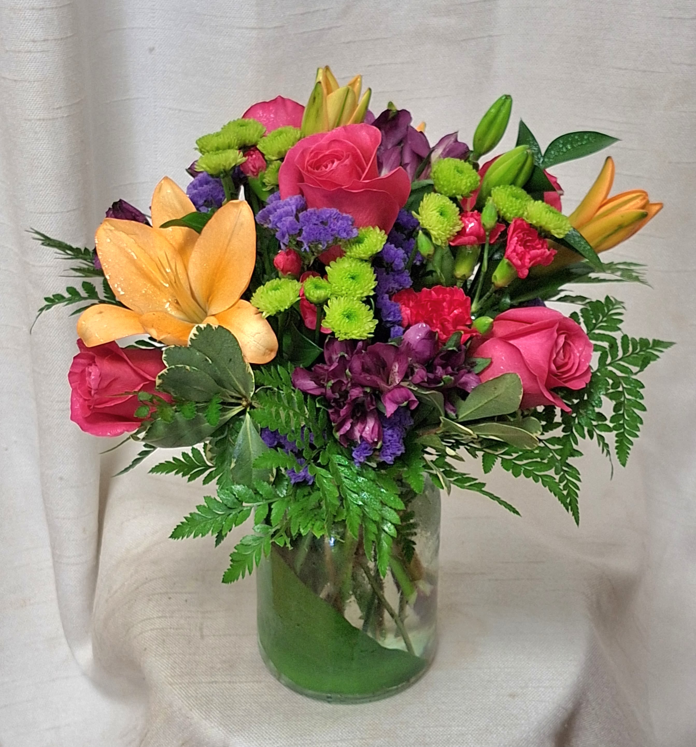 Picnic Perfection - Introducing a stunning and vibrant floral arrangement that will add a burst of color and energy to any space. This arrangement features an exquisite combination of hot pink carnations, orange lilies, alstroemerias, hot pink roses, purple statice, green button poms, and purple alstroemeria, all expertly arranged in a clear, leaf-lined glass vase. 