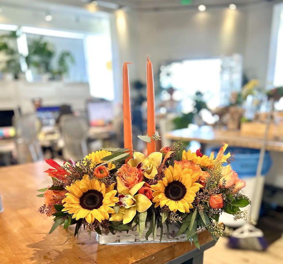 Composition with Candles - This floral centrepiece is guaranteed to create a warm and inviting atmosphere! It features radiant sunflowers, delicate yellow orchids, vibrant orange roses, and charming ranunculus, all accented by lush greenery and rustic elements. And it's finished with two elegant orange candles! It's perfect for cosy dinners or festive celebrations.