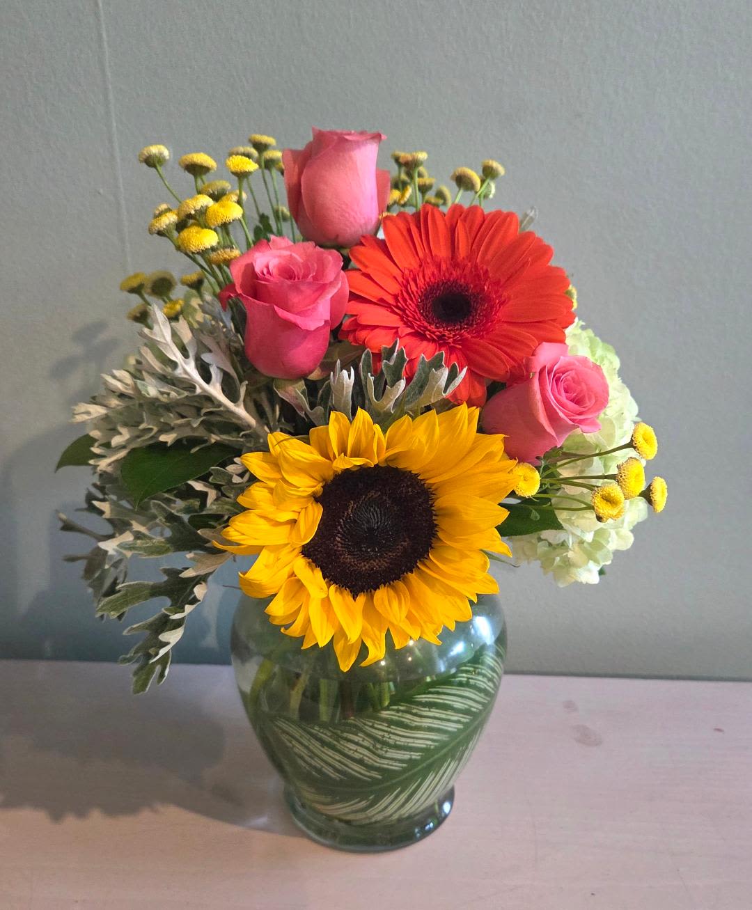 Summer Garden Symphony - Bright and beautiful summer blooms with several locally sourced flowers. Including Sunflower, Roses, Hydrangea, Gerberas accented with lemon leaf and silver dusty miller. Gerbera colors may vary.