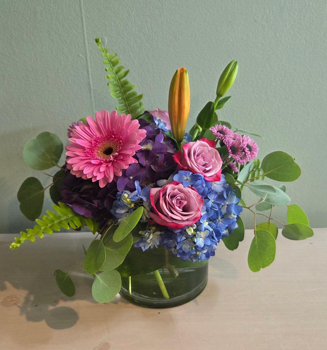Blue Belle Blue Belle - This gorgeous array of blue and purples with jewel tone accents will wow anyone and is perfect for any occasion!! Arranged in a larger 6 inch glass cylinder vase its full and a definite attention getter.