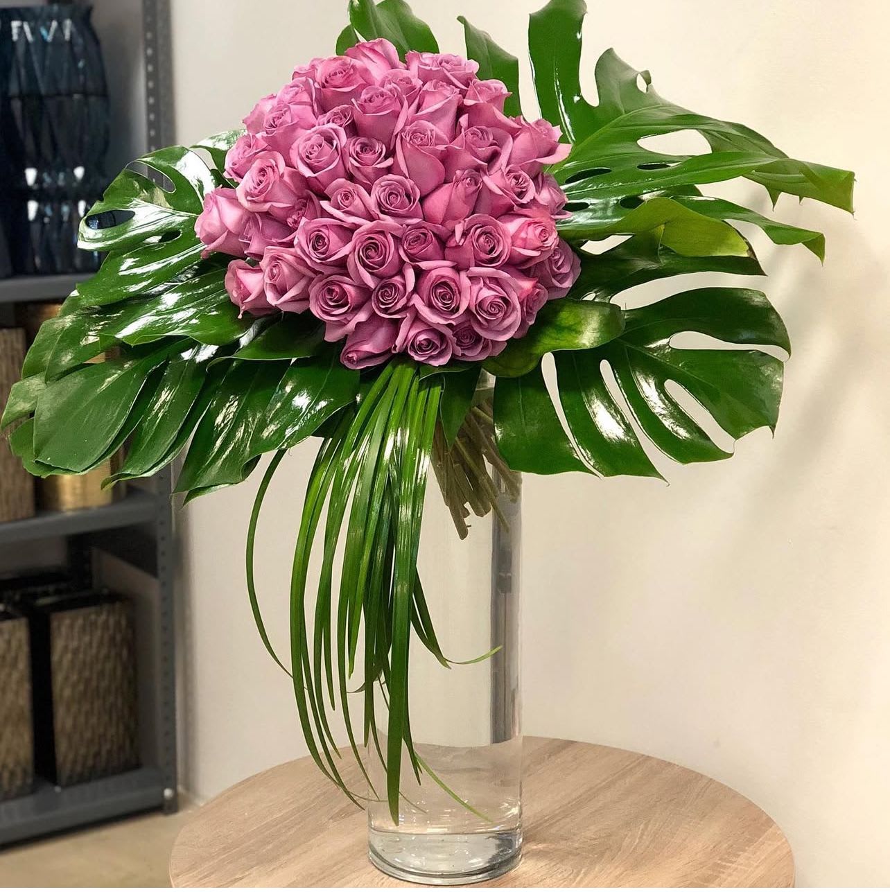 Tropical Twilight - &quot;Tropical Twilight&quot; evokes the lush beauty of a tropical sunset.  Deep purple roses are artfully arranged alongside dramatic monstera leaves, creating a striking and unforgettable display. The combination of rich color and exotic foliage makes this arrangement a captivating statement piece.  Approximately 40 roses in the standard size